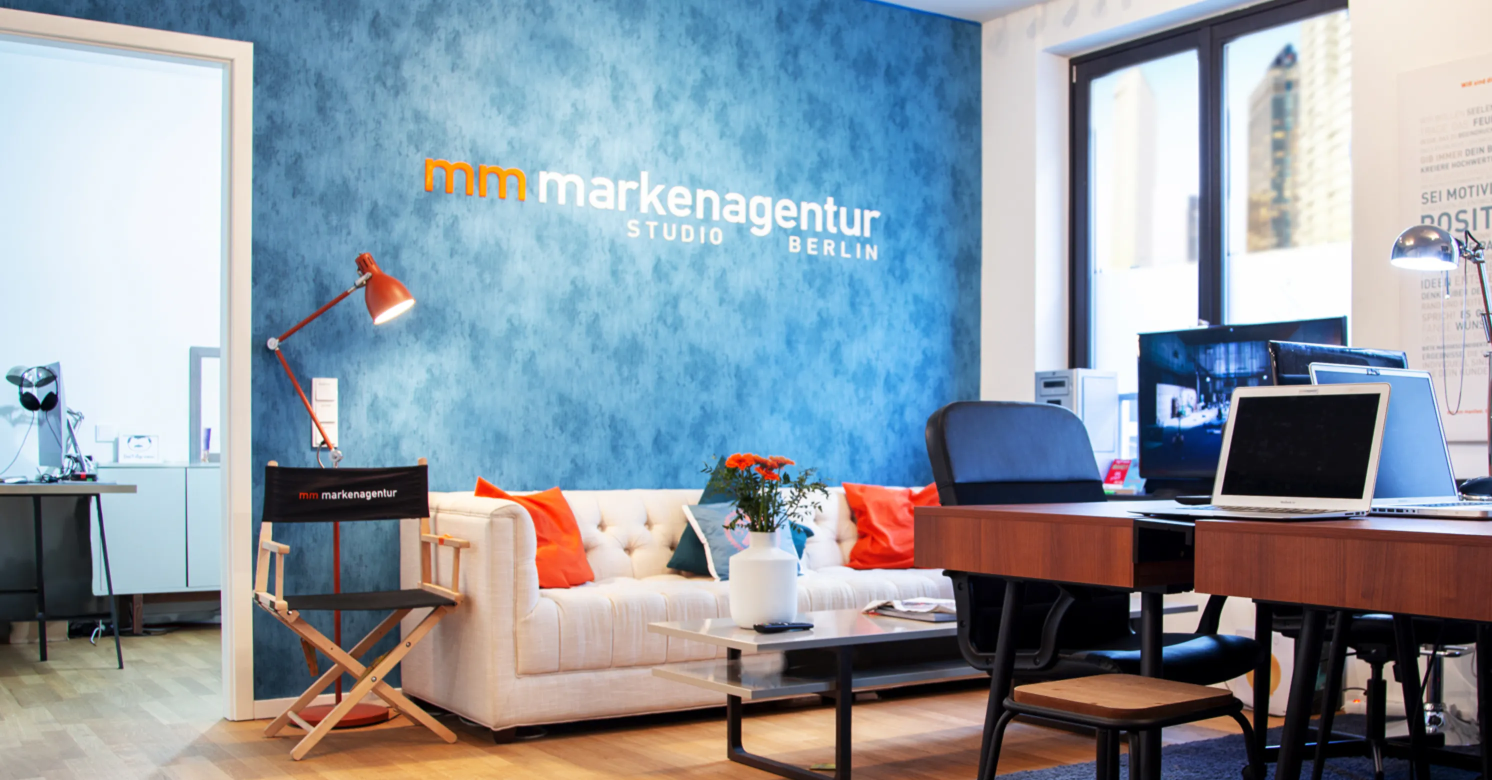 Büro mit blauer Wand, Sofa mit bunten Kissen, Tisch mit Vase und Blumen, Laptop auf Holztisch, schwarzer Stuhl, Regiestuhl und große Fenster mit Stadtblick.