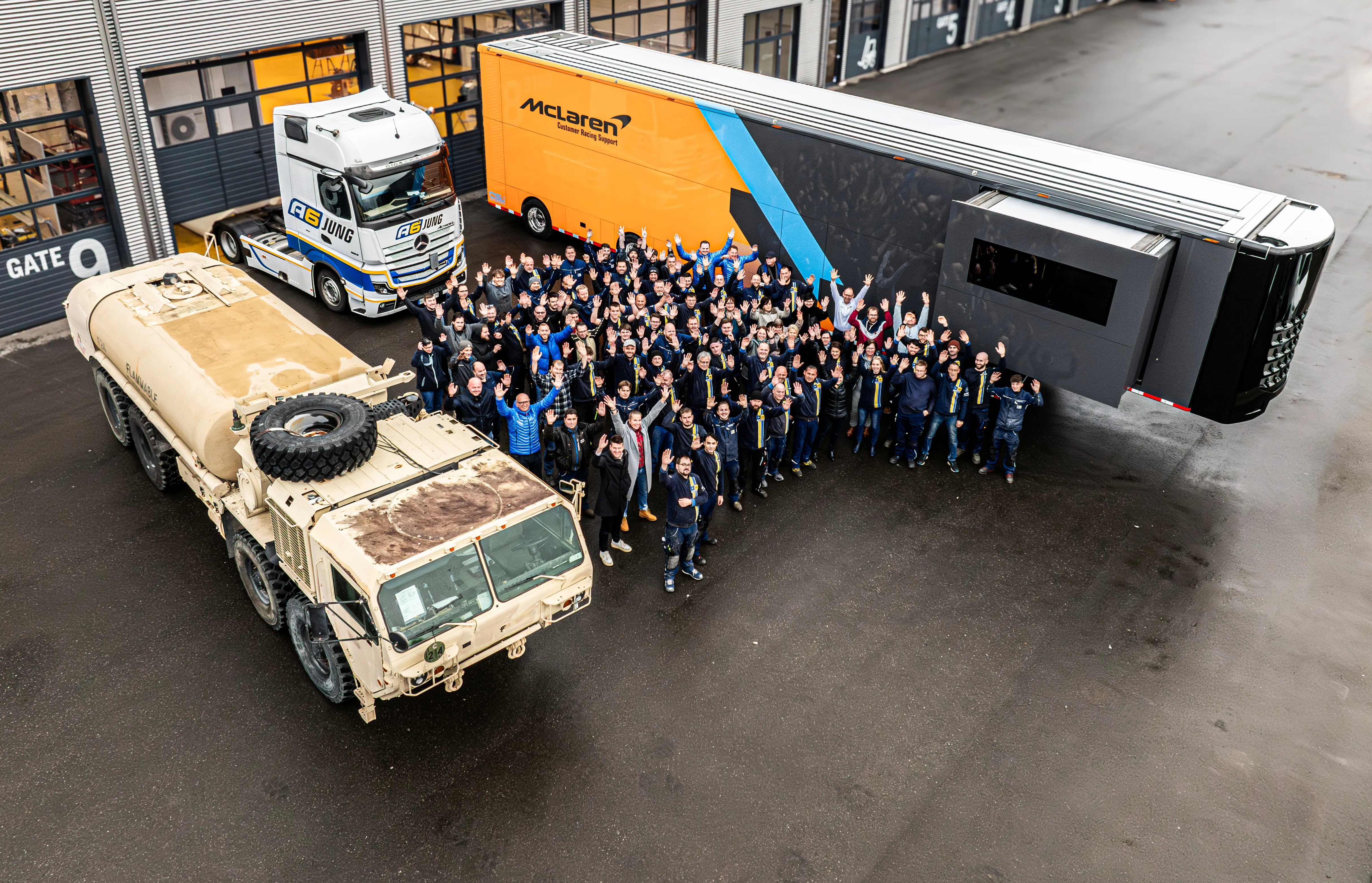 Große Gruppe von Menschen winkt zwischen einem gelb-schwarzen McLaren-Rennsupport-Lkw und einem beige Militärfahrzeug auf einem Parkplatz vor einem Gebäude.