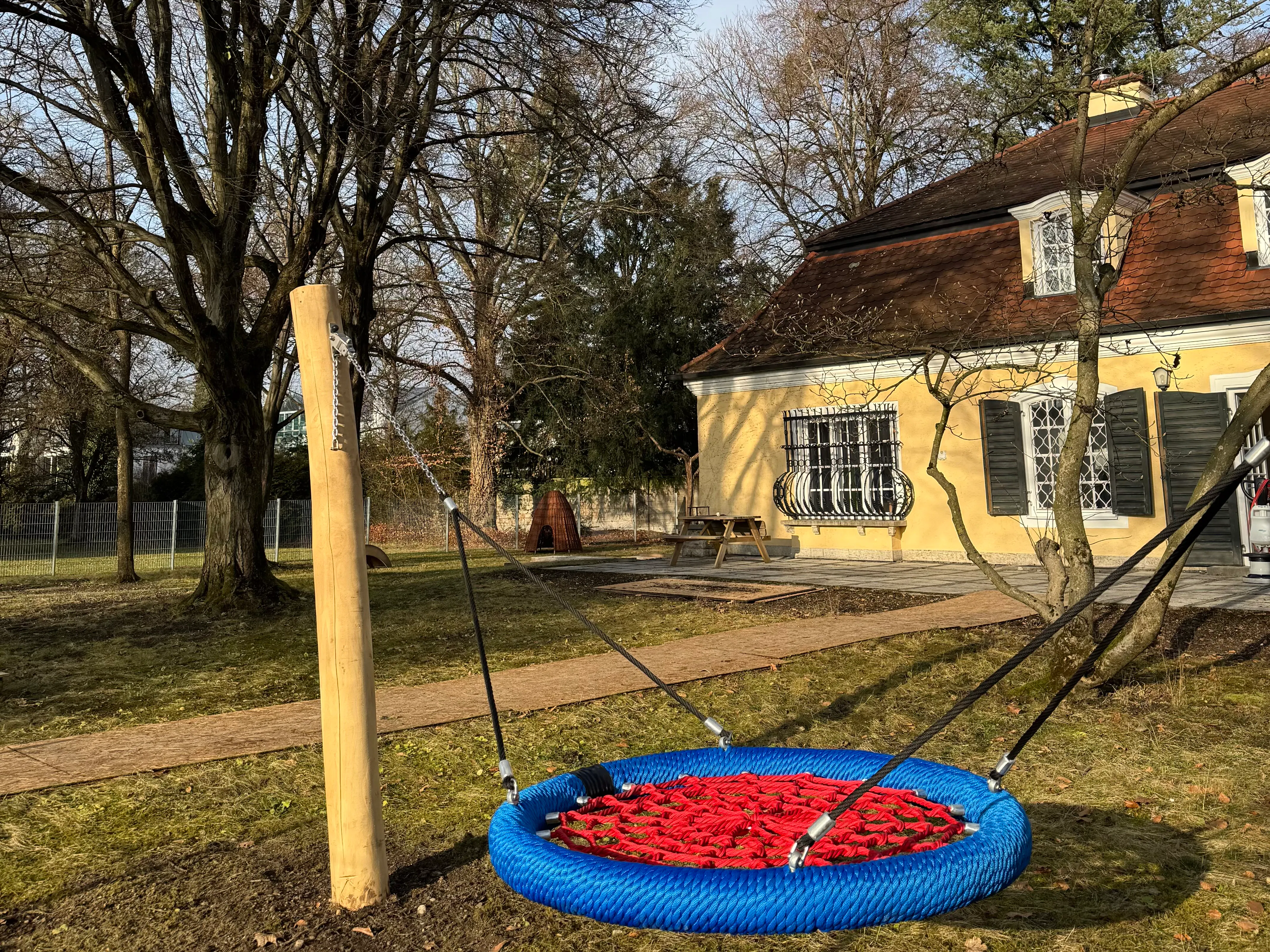 Schaukel mit rundem blauen Rahmen und rotem Netz, an drei Seilen befestigt, vor einem gelben Haus mit grünen Fensterläden und kahlen Bäumen auf einer Rasenfläche.