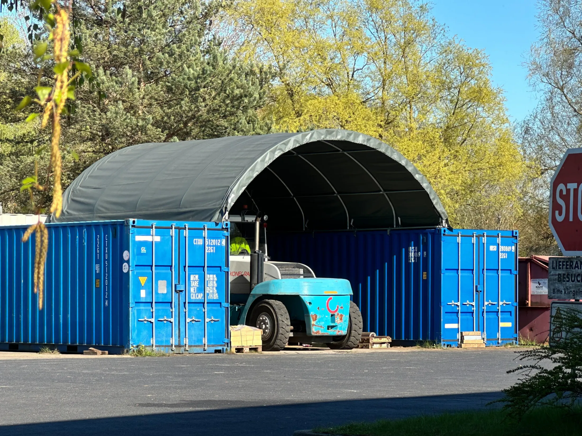 Blauer Gabelstapler steht zwischen zwei blauen Container mit grauem Zeltdach, umgeben von grünen Bäumen, rechter Bildrand zeigt teilweise STOP-Schild und Schilder mit deutscher Beschriftung.