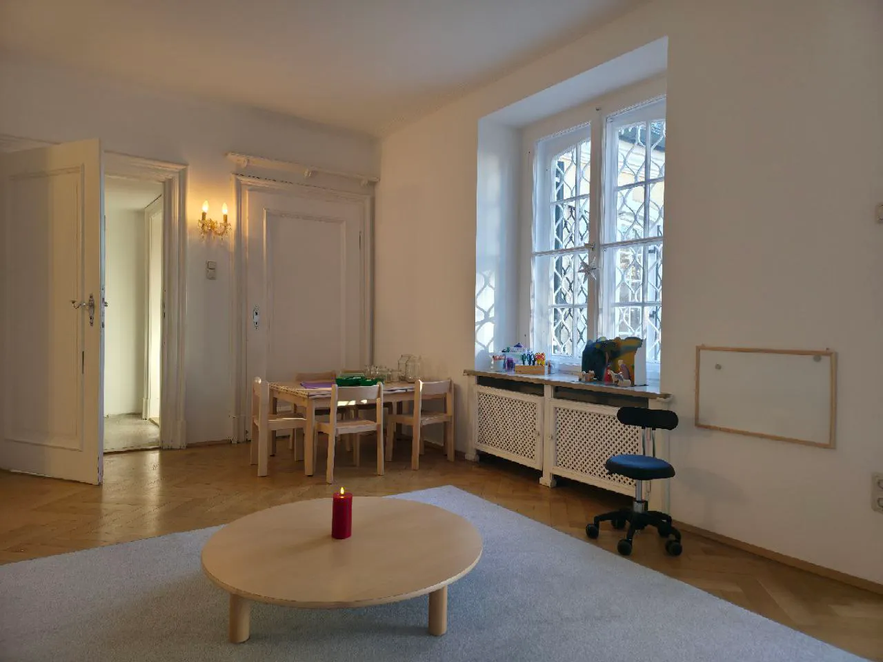 Runder niedriger Holztisch mit roter Kerze auf grauem Teppich, kleiner Holzkindertisch mit vier Stühlen, Fenster mit Gitter, schwarzer Drehstuhl und weiße Wände in hellem Raum.