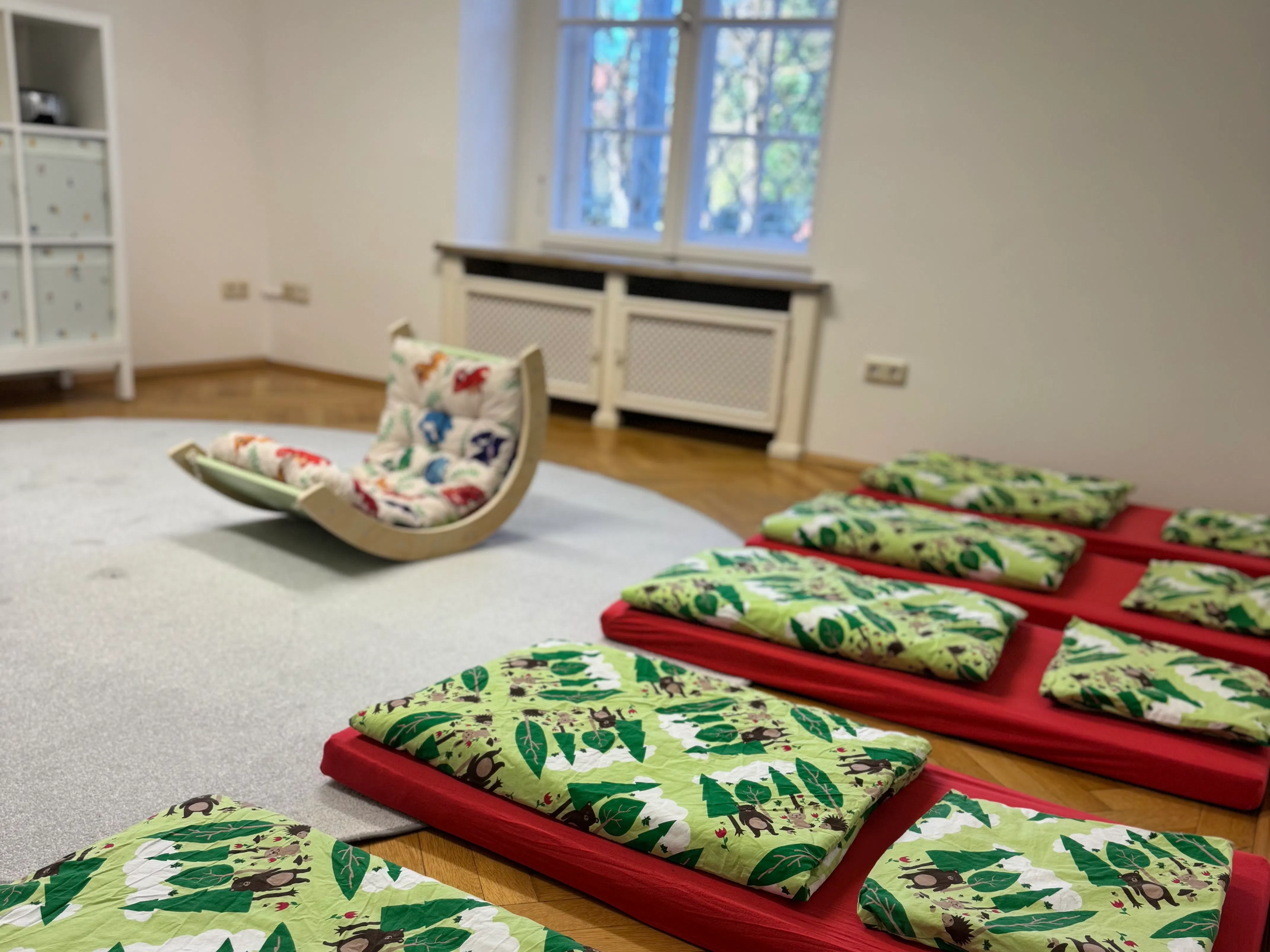 Kleine Matratzen mit grünem Blatt- und Tiermuster auf rotem Untergrund, daneben ein Holzschaukelstuhl mit gepolstertem Sitz mit Tiermotiven, helle Wände und Fenster im Hintergrund.