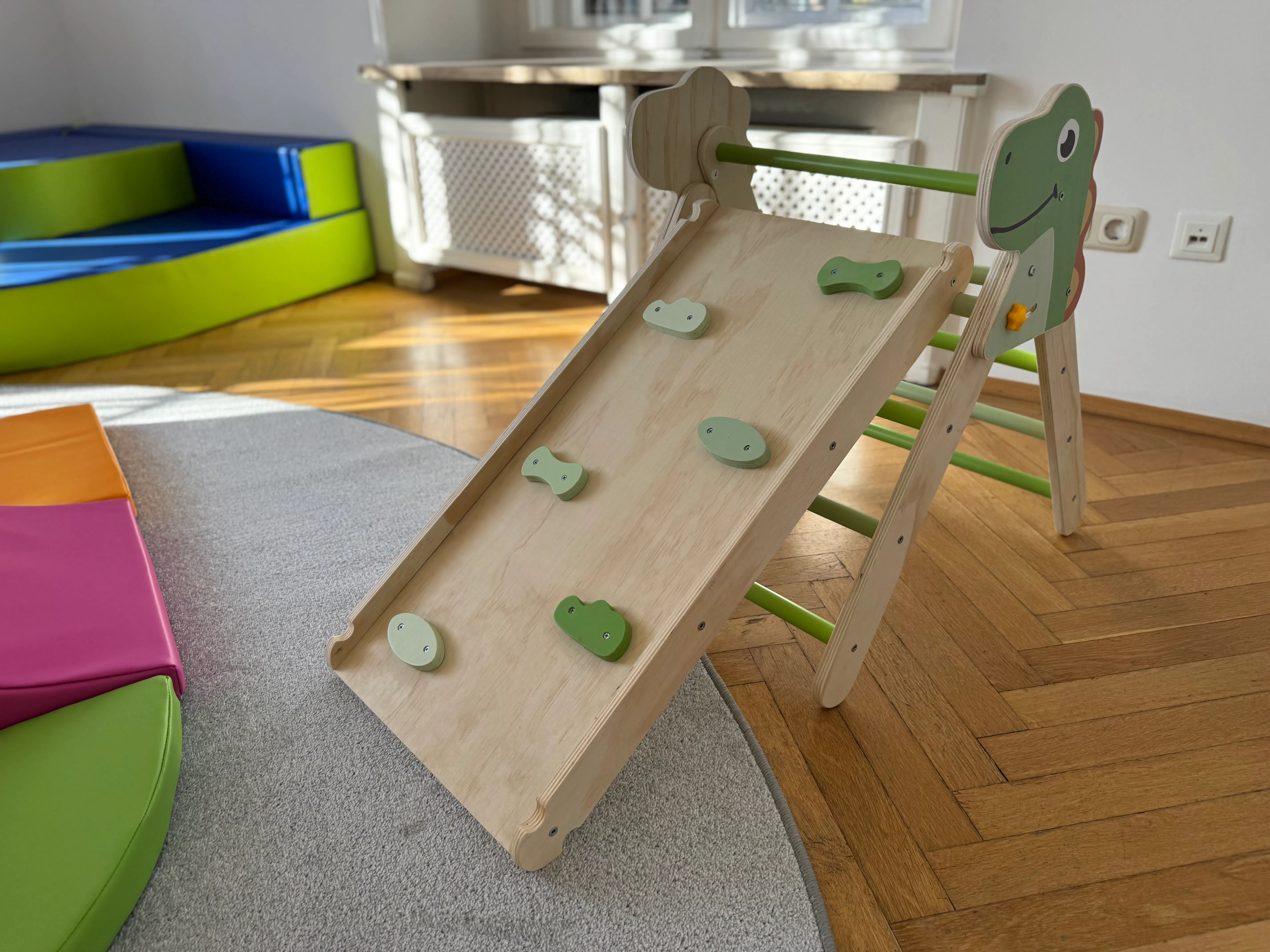 Holzspielgerät mit Kletterrampe und grünen Griffen, gestaltet wie ein Dinosaurier, auf einem Teppich in einem hellen Raum neben bunten Matten.