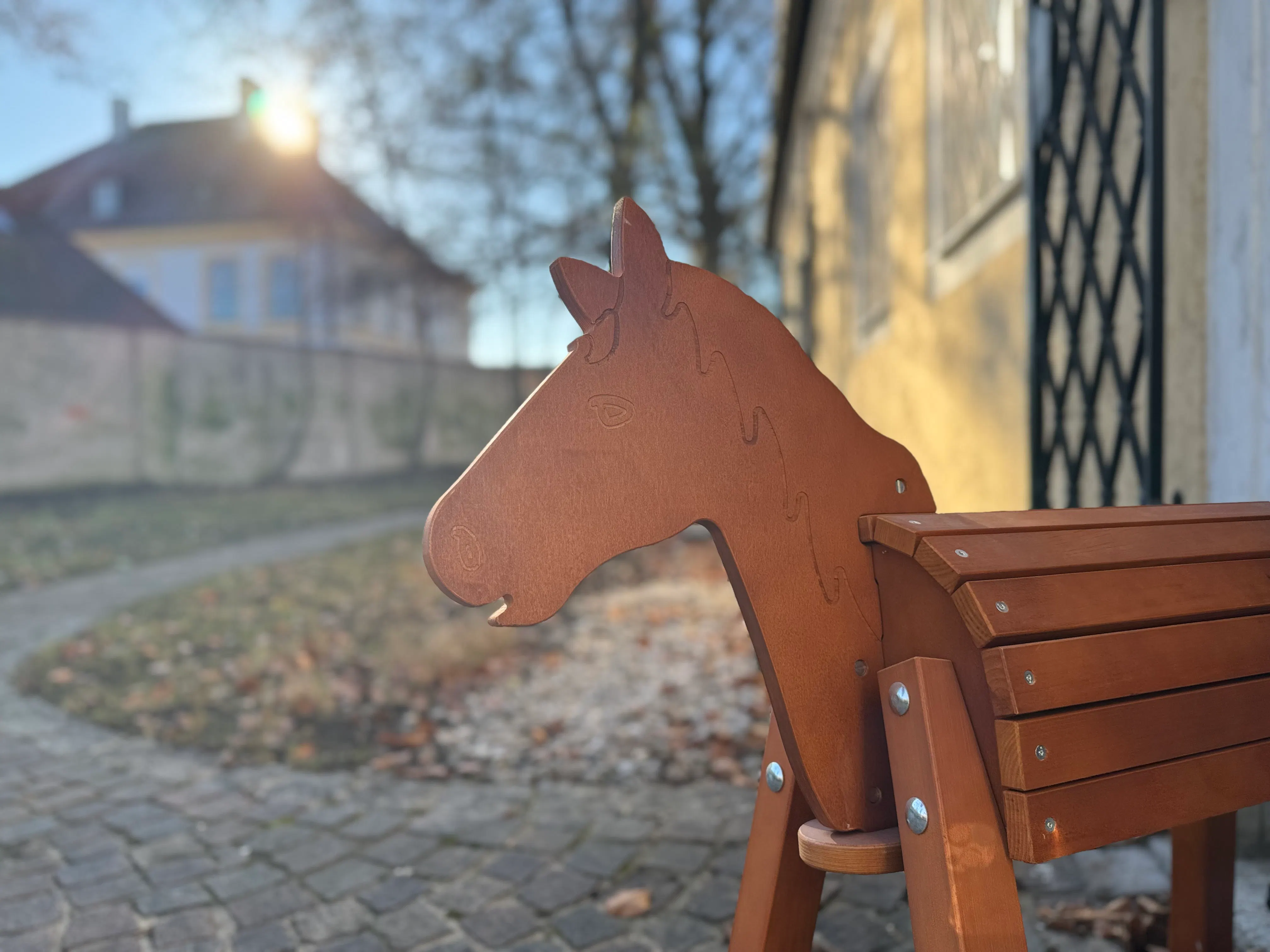 Holzbank mit Pferdekopf als Lehne, im Hintergrund unscharf ein Haus und Bäume bei Sonnenschein, gepflasterter Weg und Geländer neben der Bank sichtbar.