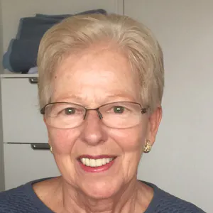 Elderly woman with short gray hair, glasses, and gold earrings, smiling and wearing a blue top, indoor background with a white cabinet and blue bag.