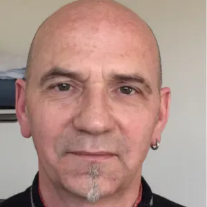 Bald man with a small gray goatee and a hoop earring in his left ear, wearing a dark sweater, against a light neutral background.