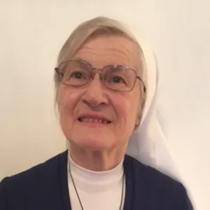 Senior woman wearing glasses and a white religious habit head covering, smiling slightly against a plain light background.