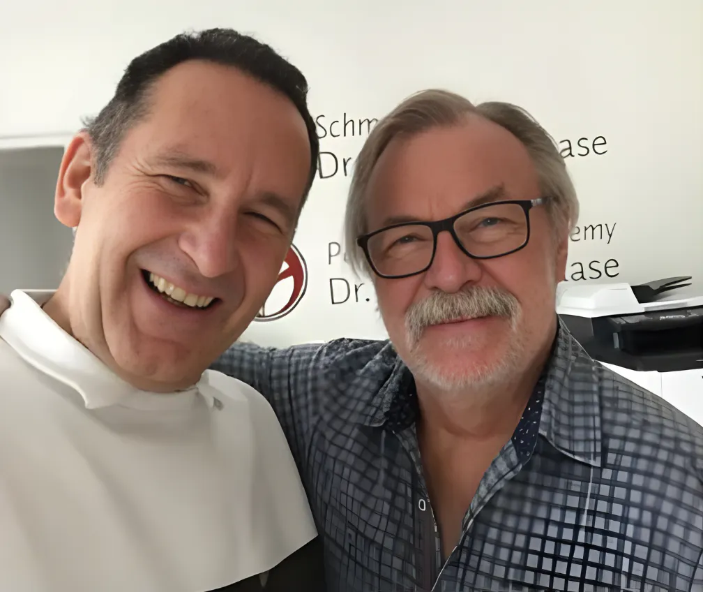 Zwei lächelnde Männer, einer trägt ein weißes Oberteil, der andere ein kariertes Hemd und Brille, vor einer Wand mit teilweise sichtbarem schwarzem Text.