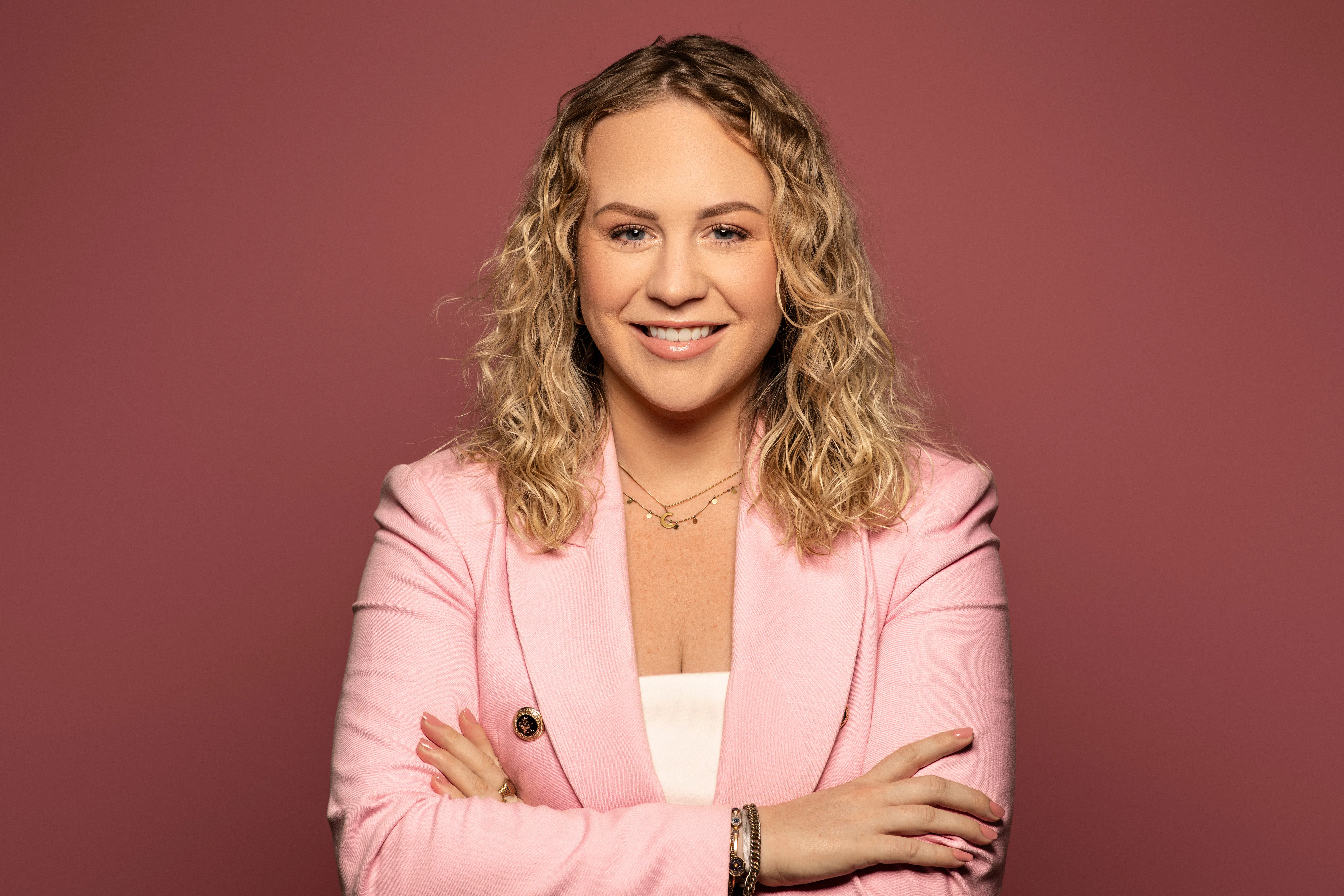 Blonde woman with curly hair smiling, wearing a pink blazer and white top, arms crossed, standing against a solid dusty rose background.