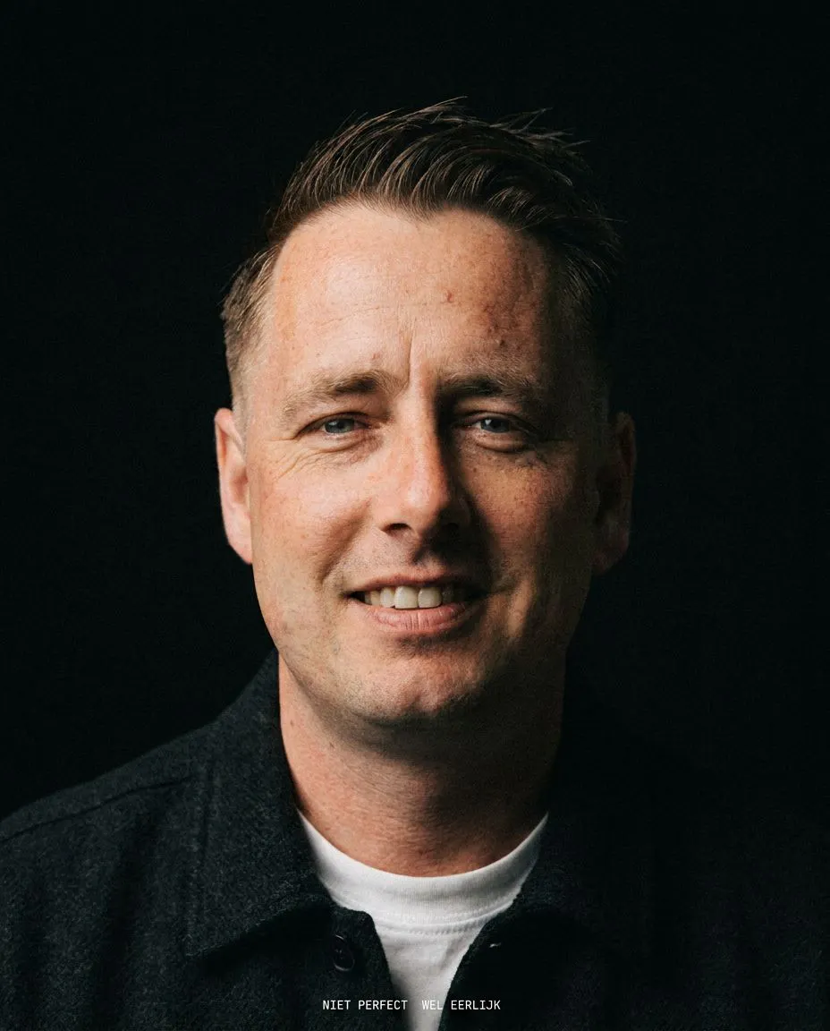 Man with short brown hair and blue eyes, smiling slightly, wearing a dark jacket over a white shirt against a black background with the text "NIET PERFECT WEL EERLIJK".