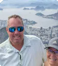 Two people smiling with a cityscape and coastal view in the background, including water, buildings, and distant mountains.