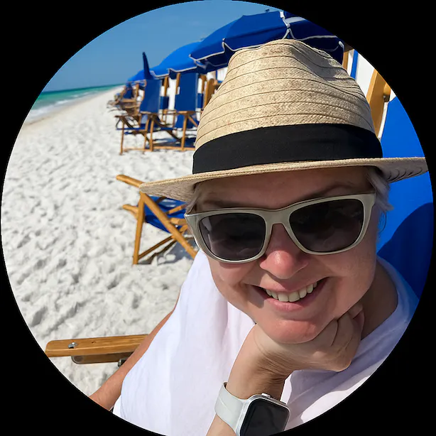 Person wearing sunglasses and a woven hat smiling on a beach with blue umbrellas and wooden chairs along the white sandy shore.