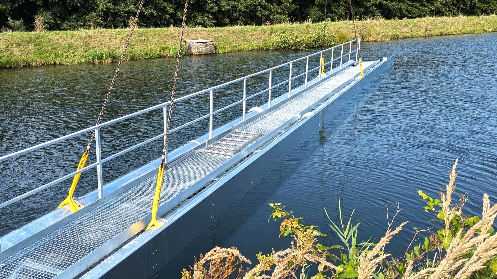 Metallgangway mit Geländer, die über einen Kanal oder Teich führt, umgeben von Gras und Pflanzen.