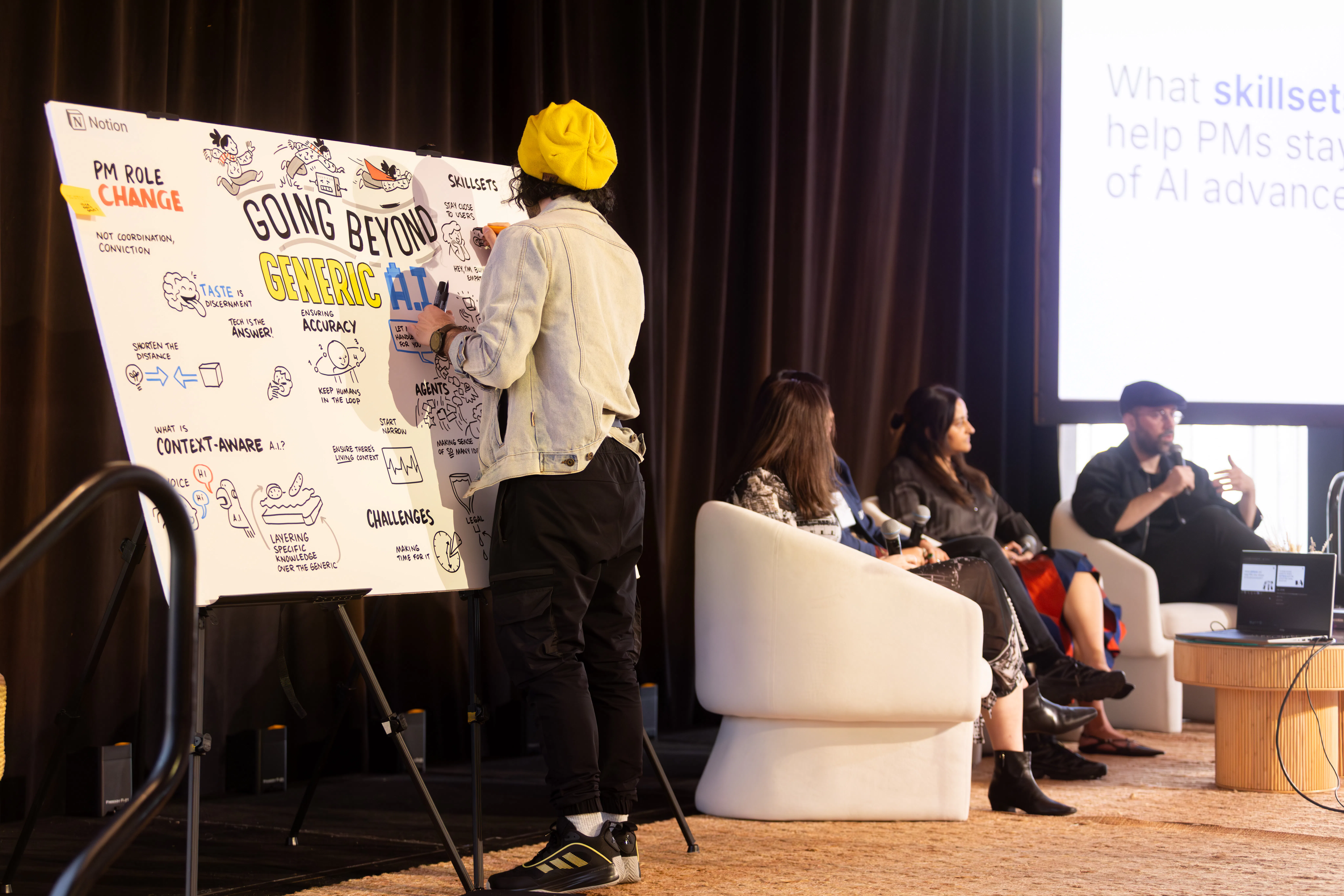 Person wearing a yellow beanie and light jacket drawing on a large board with text "Going Beyond Generic A.I." while three people seated on white chairs hold microphones in the background.