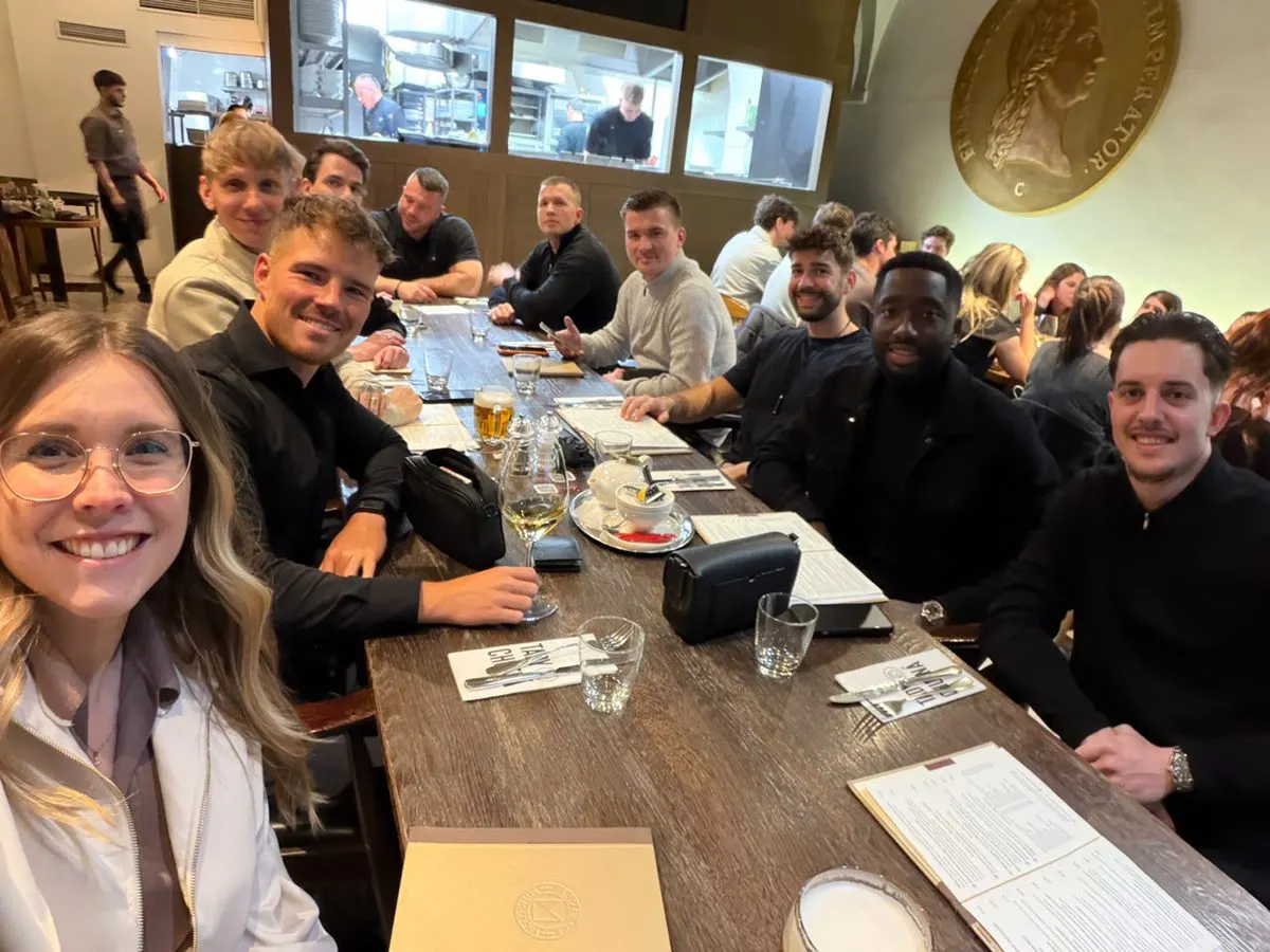 Group of twelve young adults sitting around a long wooden table in a restaurant, with menus, glasses, and drinks, smiling at the camera.