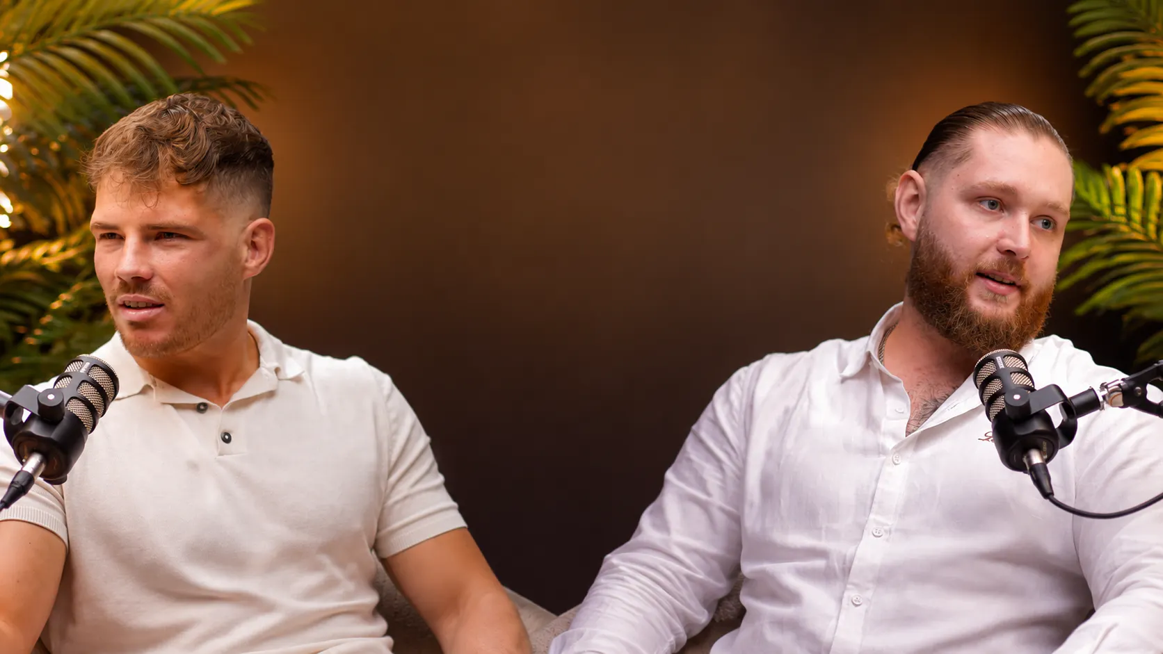 Zwei Männer mit Mikrofonen sitzen nebeneinander, einer trägt ein weißes Polohemd, der andere ein weißes Hemd, und führen ein Gespräch in einem Studio mit Pflanzen im Hintergrund.