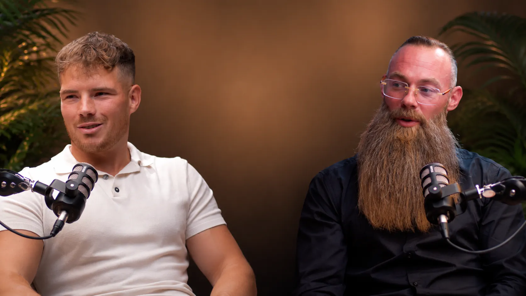 Zwei Männer mit Mikrofonen, einer trägt ein weißes Polohemd und hat kurze blonde Haare, der andere hat ein schwarzes Hemd, Brille und langen, dichten Bart.