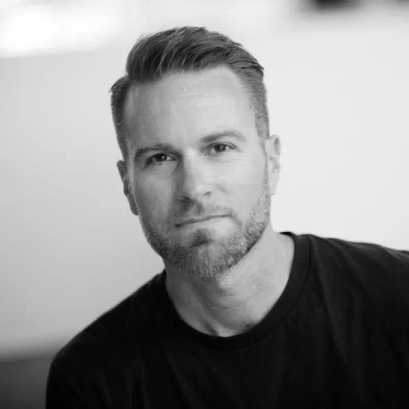 Man with short, styled hair and light beard wearing a black shirt, looking directly at the camera with a neutral expression against a blurred background.