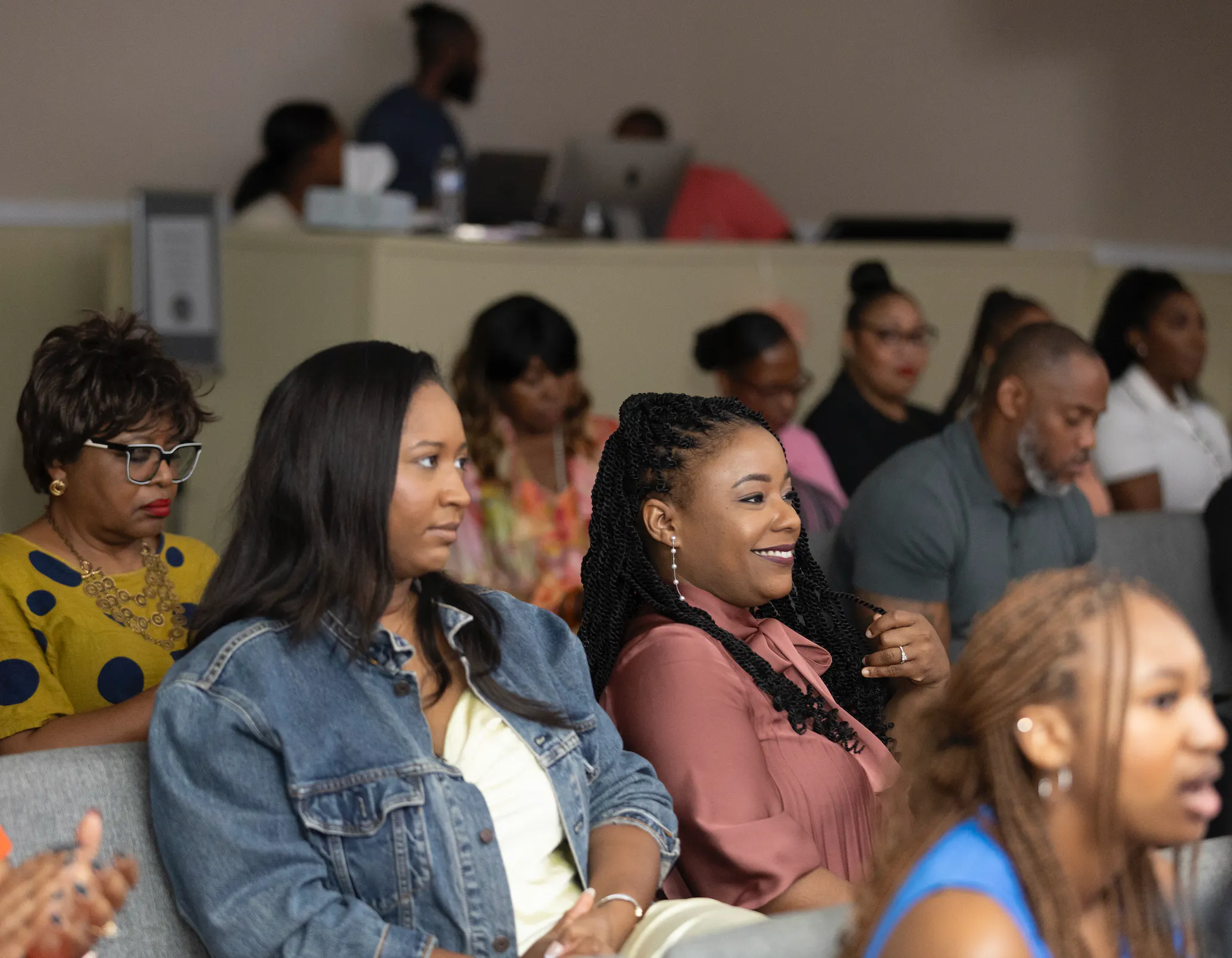 A group of diverse adults seated indoors, facing forward, some listening attentively while others appear engaged or smiling.