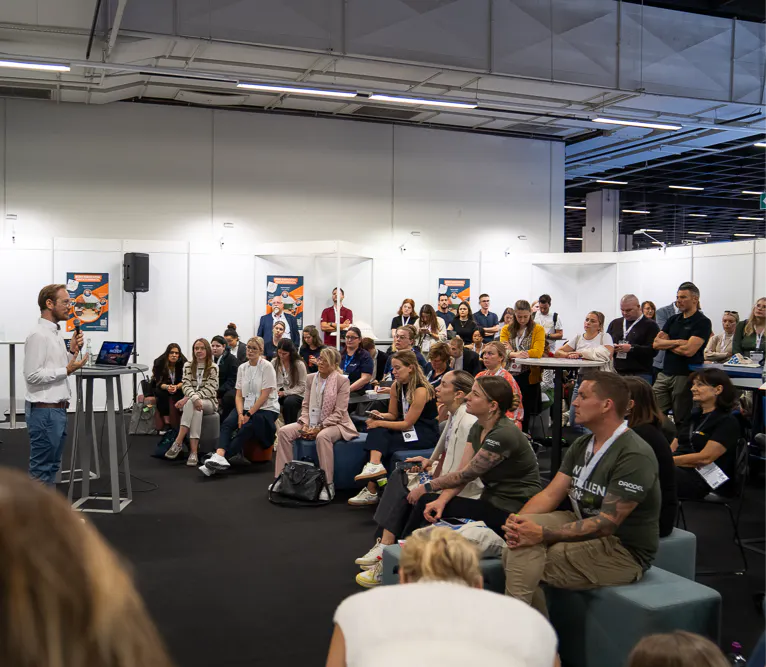 Mann hält Vortrag vor sitzendem und stehendem Publikum in einem modernen Konferenzraum mit weißen Trennwänden und Informationspostern.