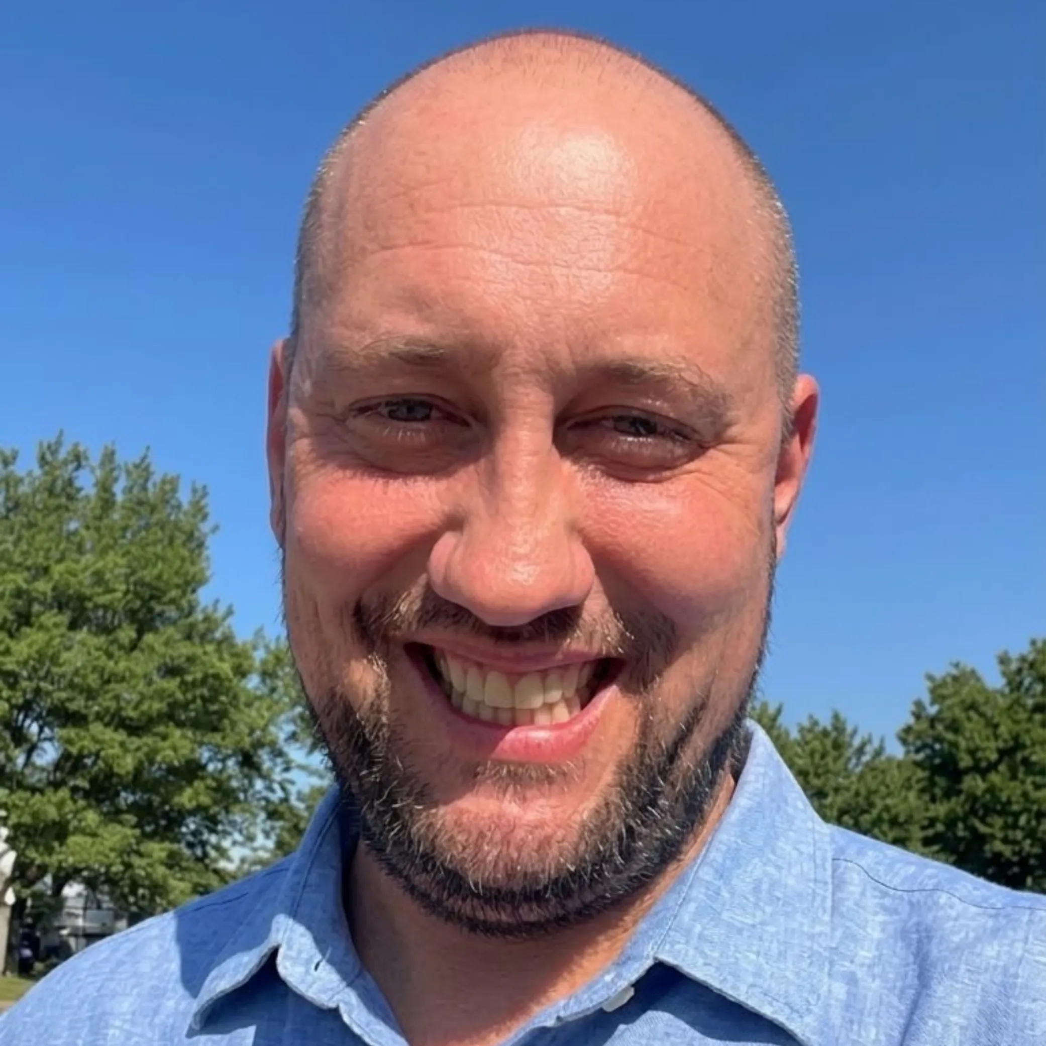 Smiling bald man with short beard wearing a blue collared shirt outdoors with green trees and clear blue sky in the background.
