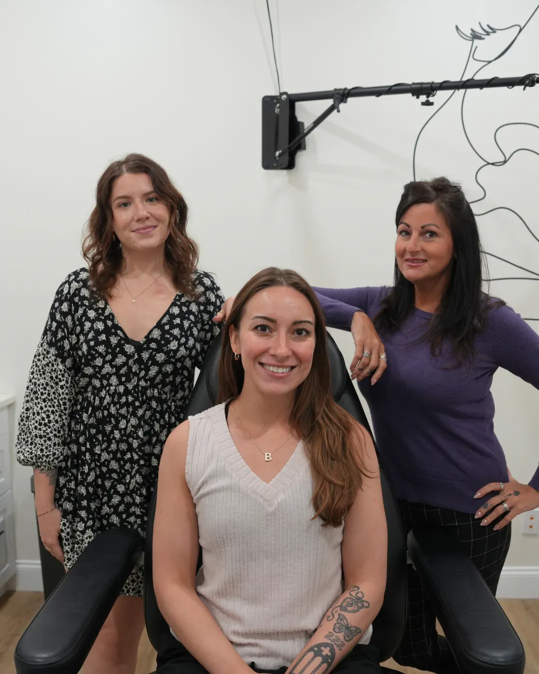 Three women smiling indoors, one seated in a chair wearing a sleeveless top with tattoos on her arm, two standing behind her with one in a black floral dress and the other in a purple sweater.