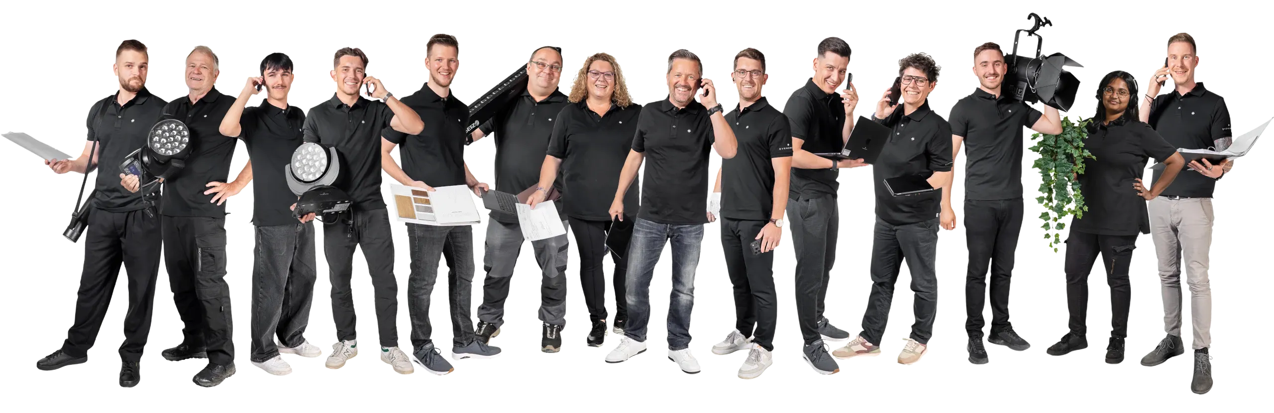Gruppe von 14 Personen in schwarzer Arbeitskleidung, einige halten technische Geräte, Akten oder Pflanzen, sie lächeln und simulieren Telefongespräche.