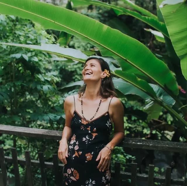 Woman wearing a black floral dress with a flower in her hair standing by a wooden railing surrounded by lush green tropical plants, smiling and looking upward.