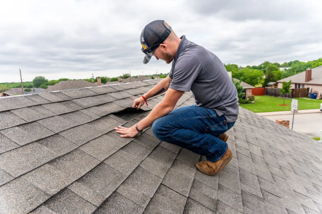 A man wearing a cap, gray polo, jeans, and brown shoes is lifting a roof shingle on a residential rooftop, inspecting it under cloudy skies.