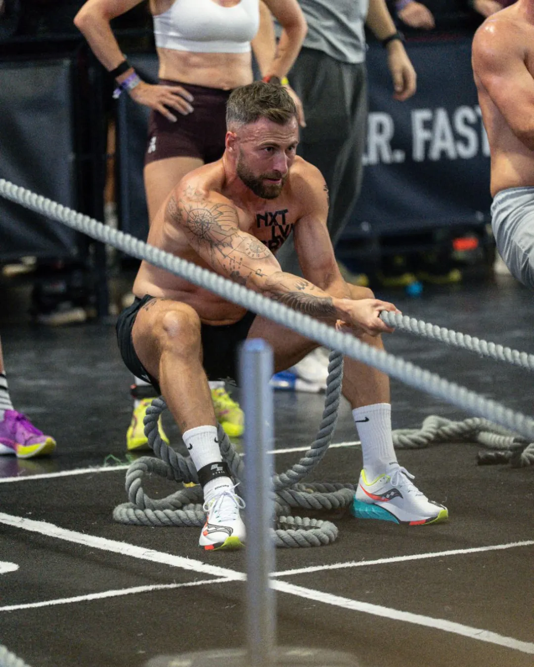 Mann ohne Oberbekleidung zieht an zwei großen Seilen in einer Sporthalle, andere Personen und Trainingsgeräte sind im Hintergrund zu sehen.