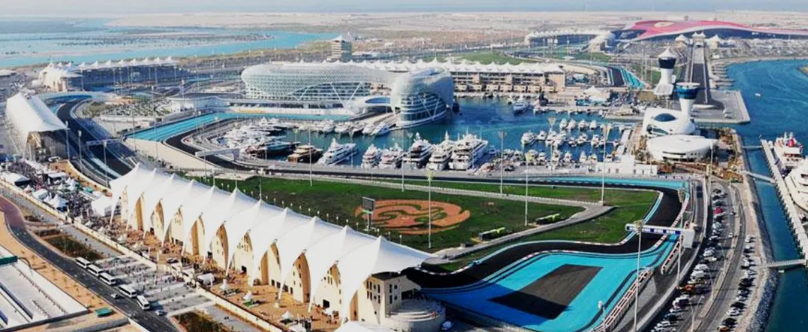 Aerial view of a marina with yachts, surrounded by modern buildings and a racing track extending around the area near a large water body.