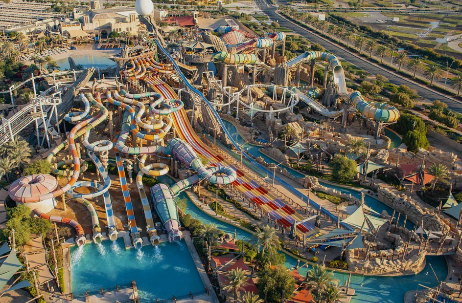 Large water park with numerous colorful water slides, pools, and winding tubes surrounded by palm trees and pathways, with a dry landscape and parking area in the background.