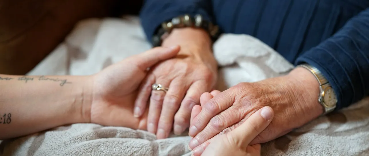 Zwei ältere Hände halten jeweils eine jüngere Hand, eine tätowierte Hand und eine weitere ältere Hand mit Schmuck und Armbanduhr liegen auf einer Decke.