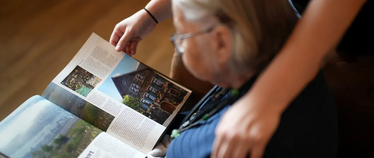 Ältere Person, die ein Magazin mit Bildern eines Hauses und Text betrachtet, während eine jüngere Person tröstend die Schulter berührt.