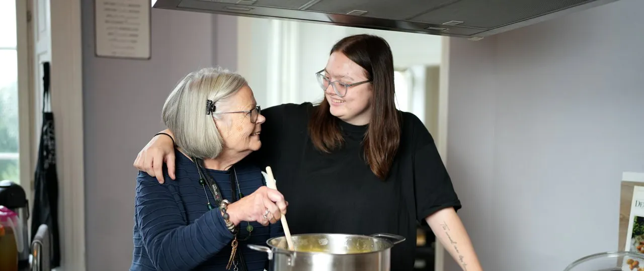 Ältere Frau und jüngere Frau schauen sich beim gemeinsamen Kochen in der Küche lächelnd an, ältere Frau rührt in einem großen Topf.