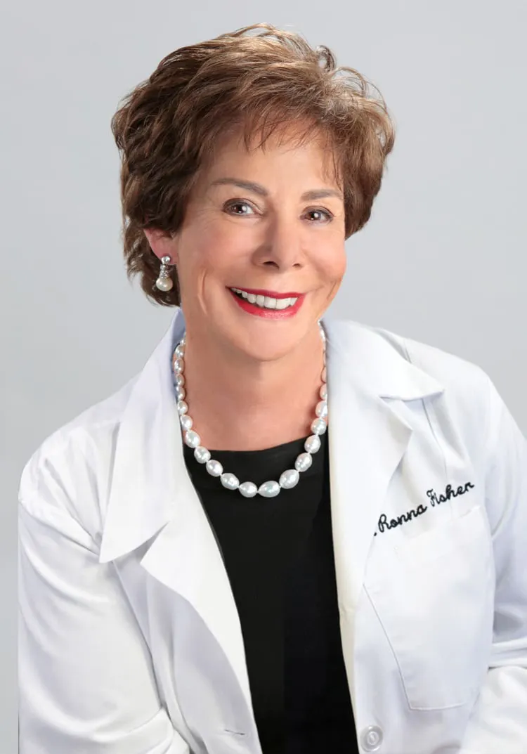 Smiling woman wearing a white coat with a pearl necklace and pearl earrings against a plain light gray background.