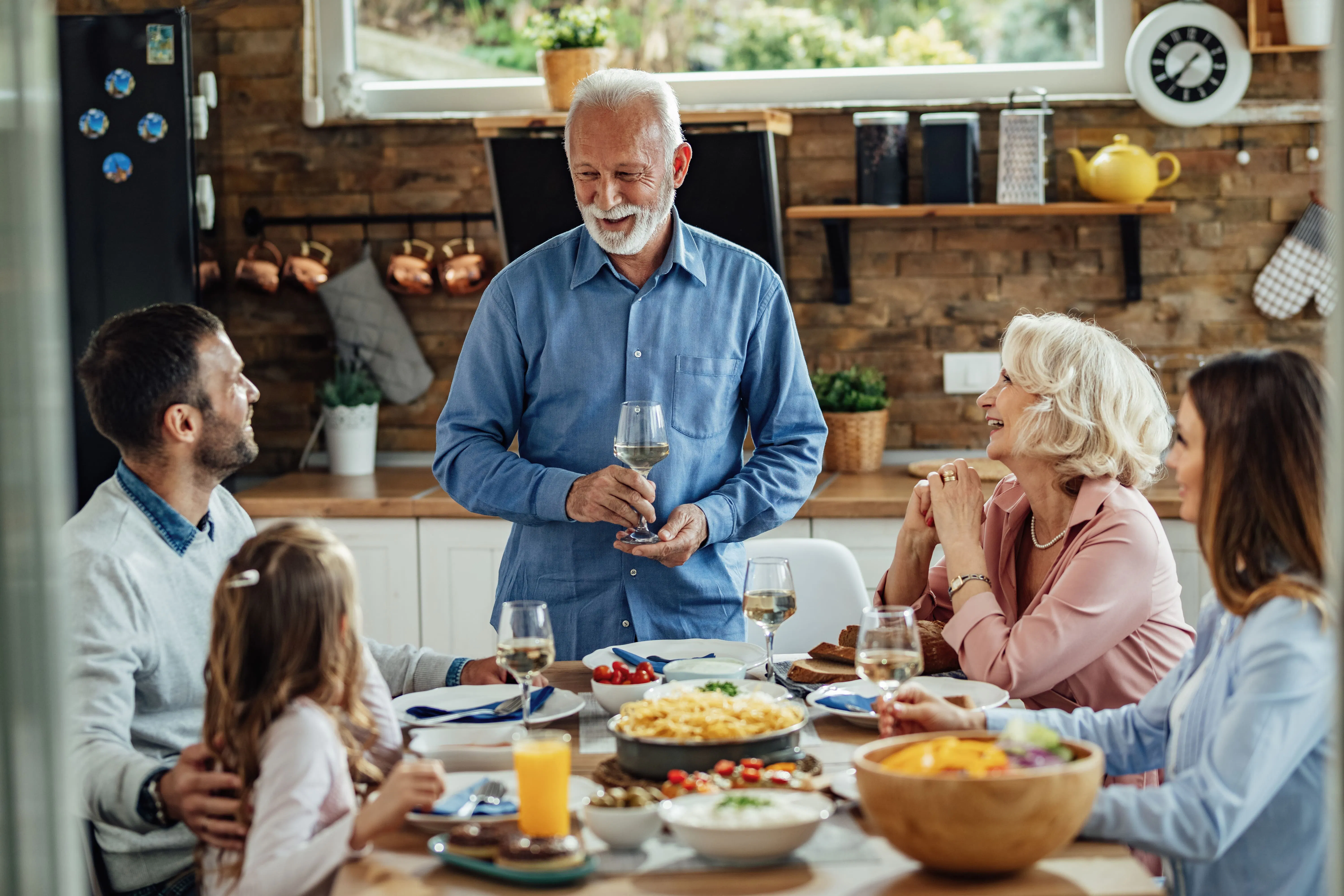 Family gathered at a kitchen table enjoying a meal with various dishes and drinks, while an older man stands holding a glass and speaking to others seated around the table.