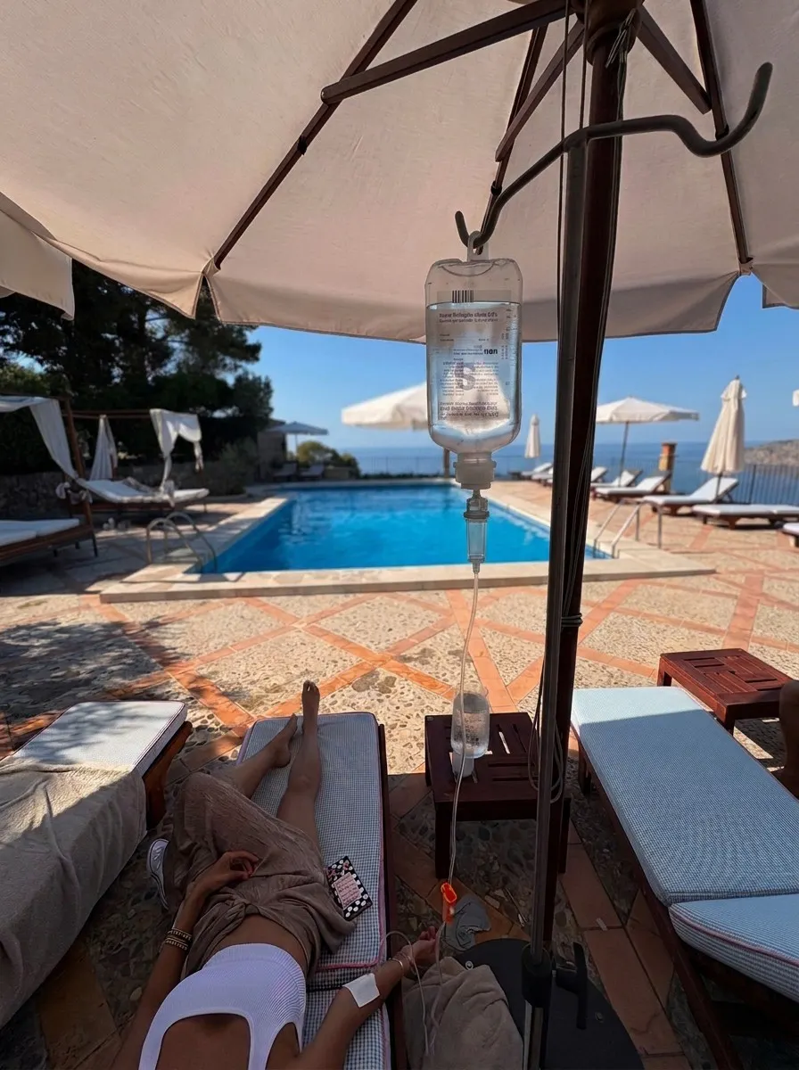Person lying on a poolside lounge chair under an umbrella with an IV drip attached to their arm, overlooking a swimming pool and sun loungers in the background.