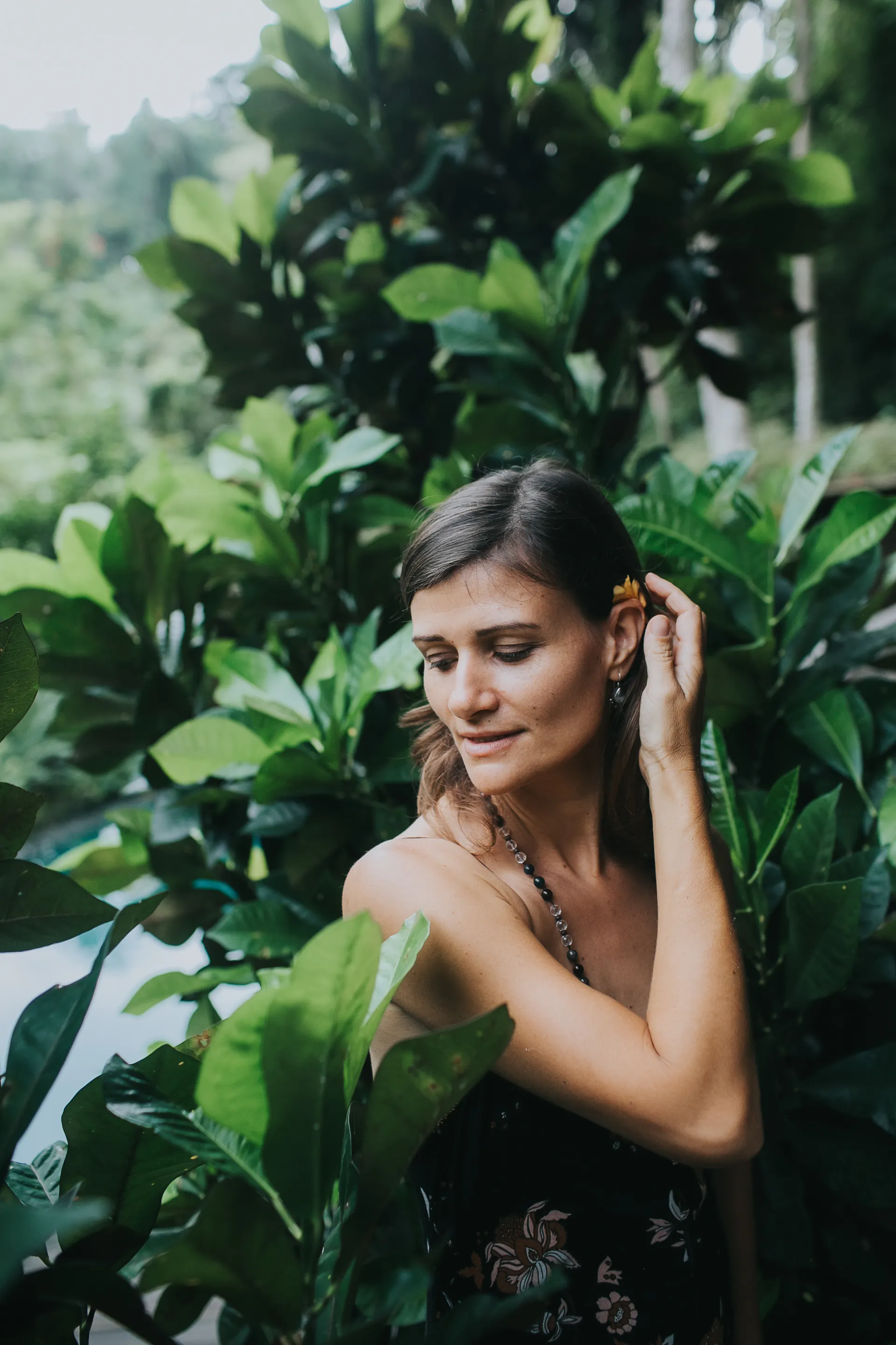 Woman with a flower in her hair standing among lush tropical green leaves, softly looking down