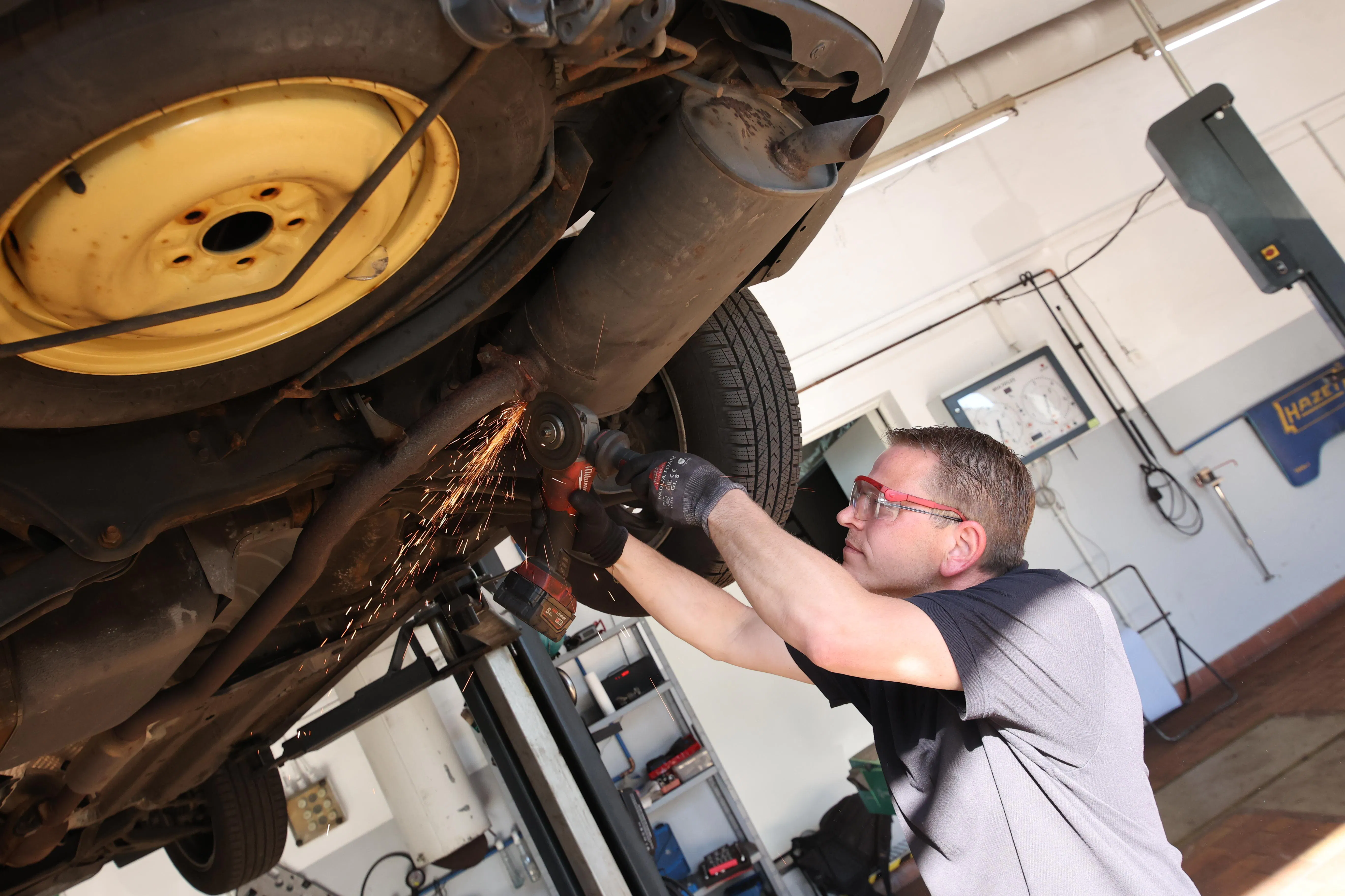 Mechanic wearing safety glasses using an angle grinder on the underside of a vehicle lifted in a garage.
