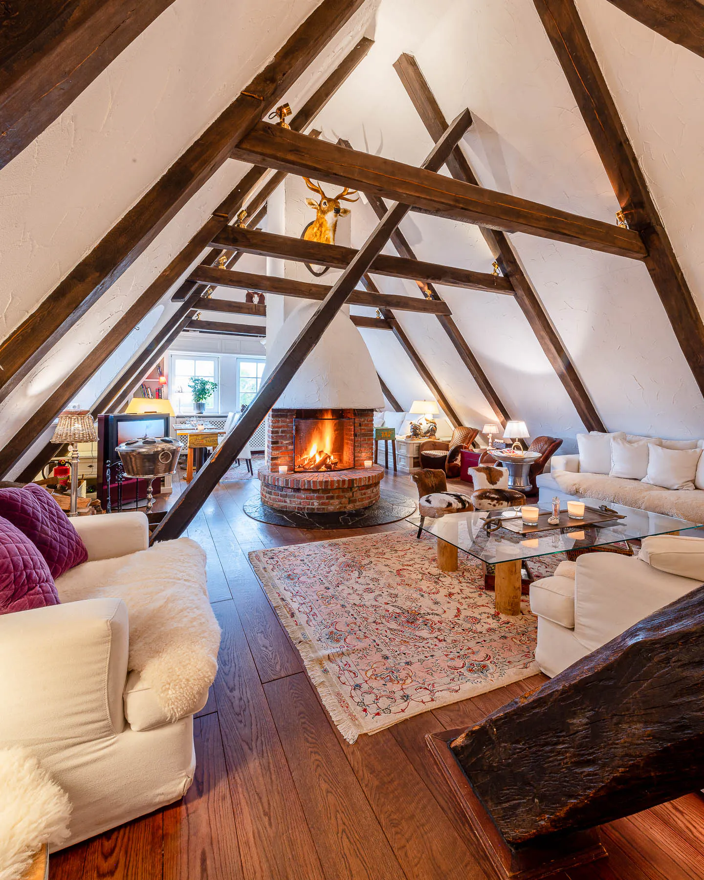 Cozy attic living room with exposed wooden beams, brick fireplace, white sofas, glass coffee table, patterned rug, and mounted deer head above the fireplace.