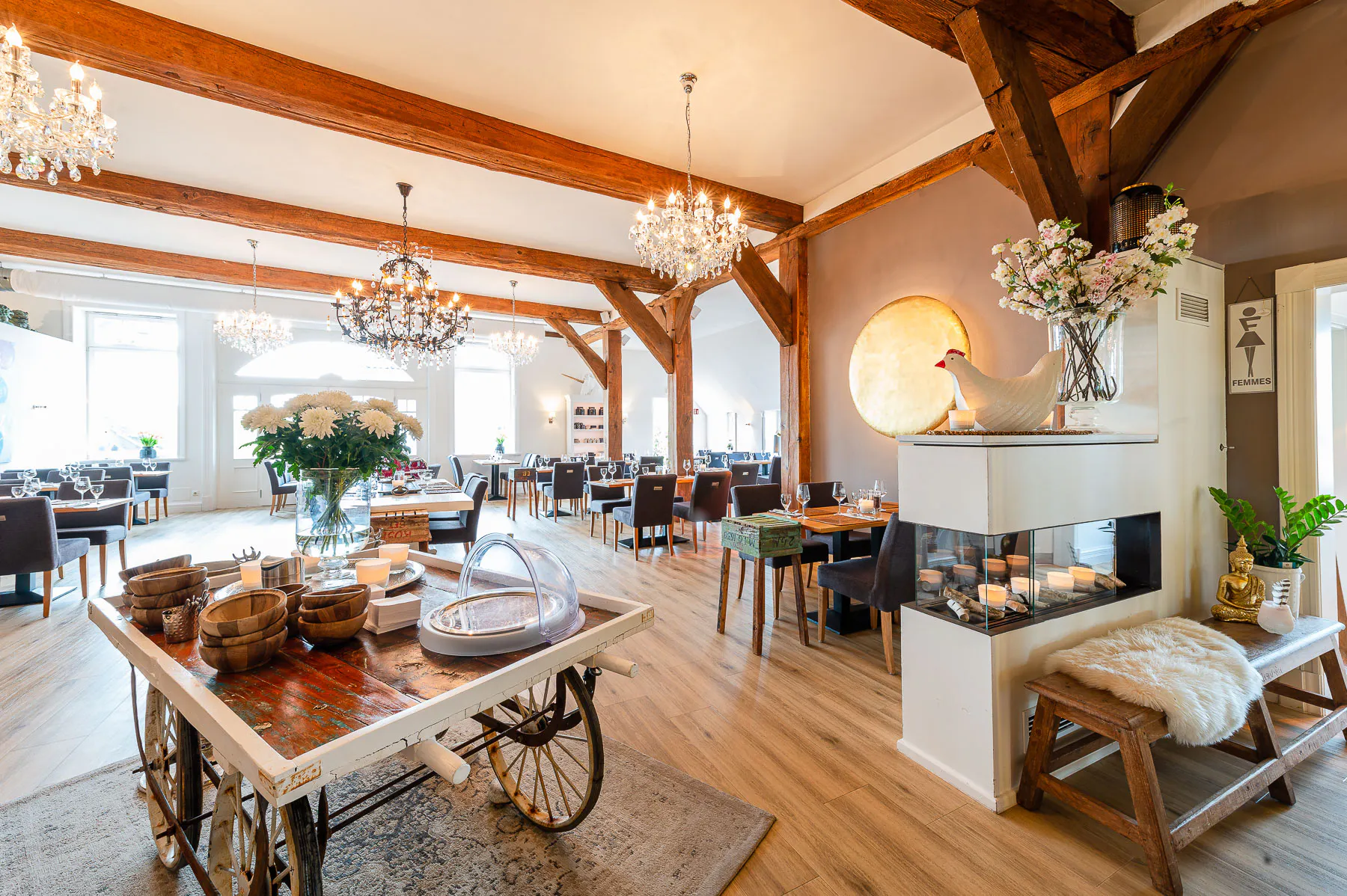 Bright restaurant interior with wooden beams, chandeliers, tables set with glasses, a decorative cart with bowls and flowers, and a cozy seating area with a faux fur throw.