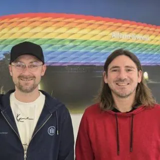Two men smiling in front of a colorful geometric wall with a rainbow pattern, one wearing a black cap and glasses, the other with long hair and a red hoodie.