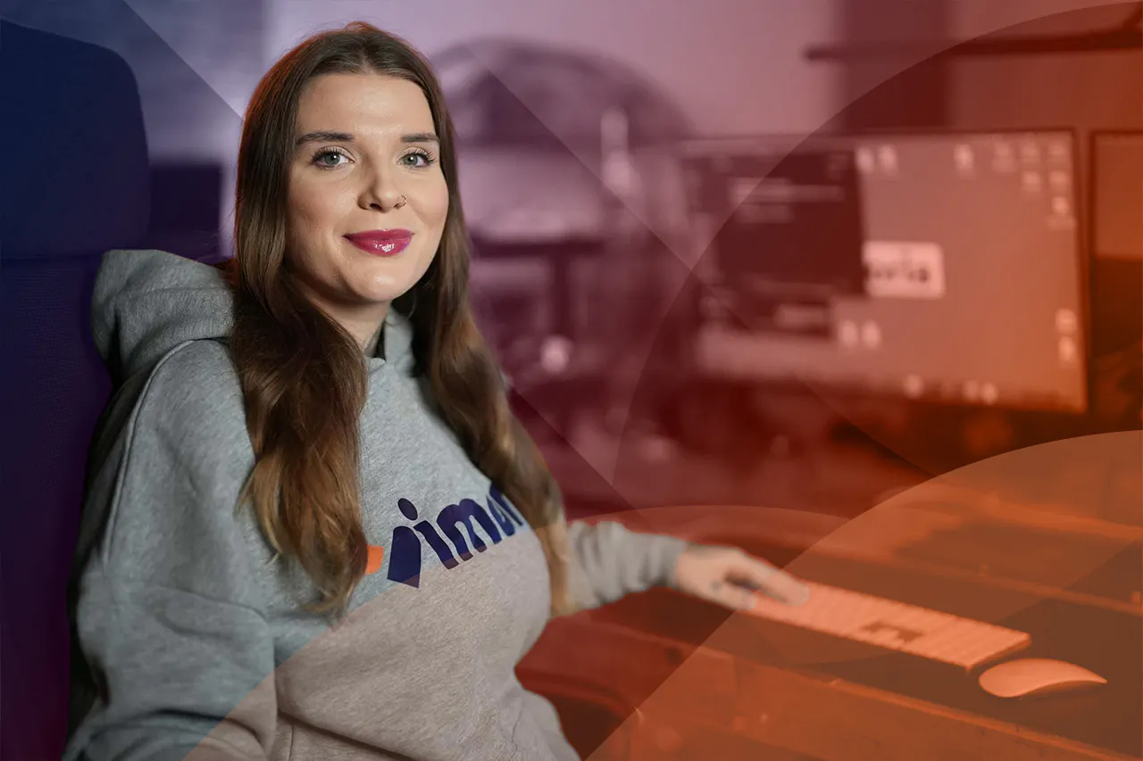 Woman with long brown hair wearing a grey hoodie sitting at a desk with a keyboard and computer monitors in the background.