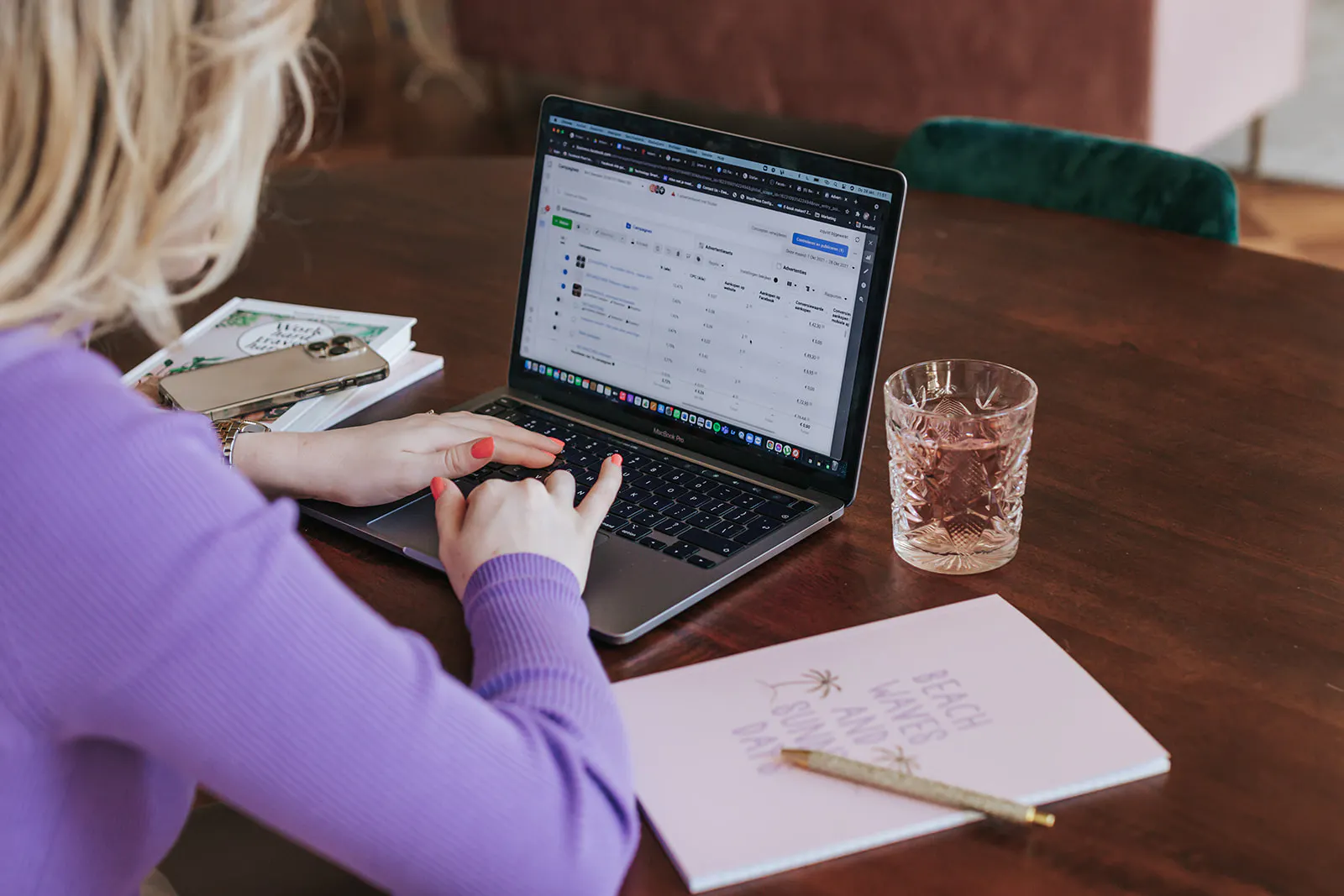 Person wearing a purple sweater typing on a MacBook Pro displaying a data table, with a notebook, pen, smartphone, and a glass of water on a wooden table.