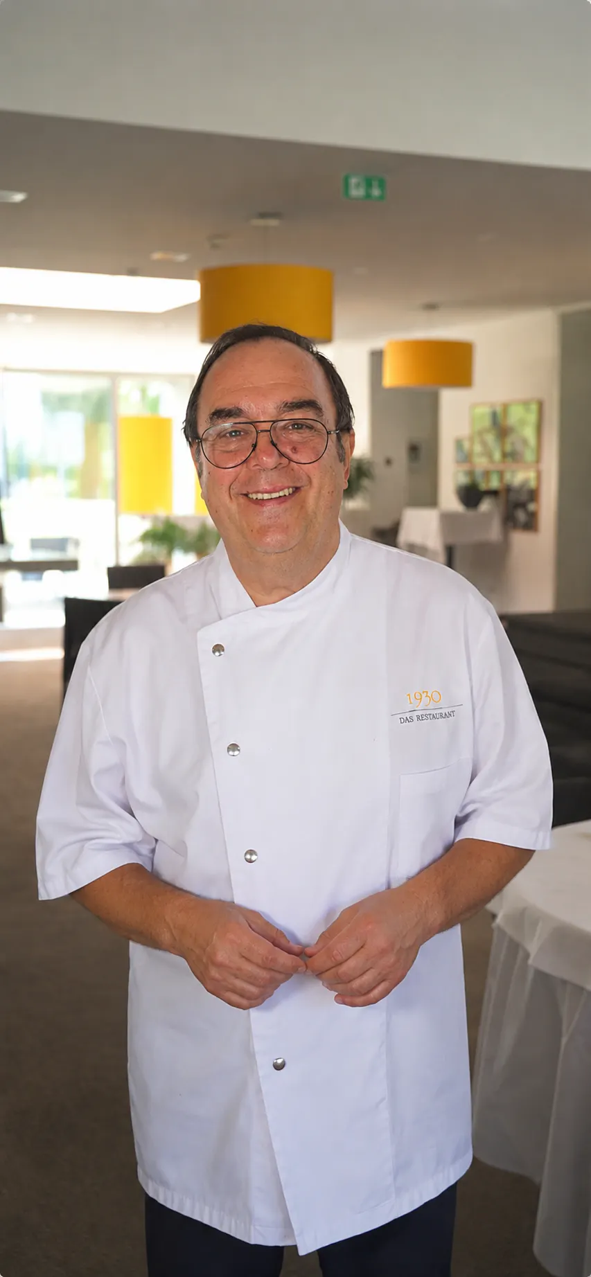 Älterer Mann mit Brille trägt weiße Kochjacke mit Aufschrift „1930 Das Restaurant“ und lächelt in einem modernen Innenraum mit gelben Lampenschirmen.