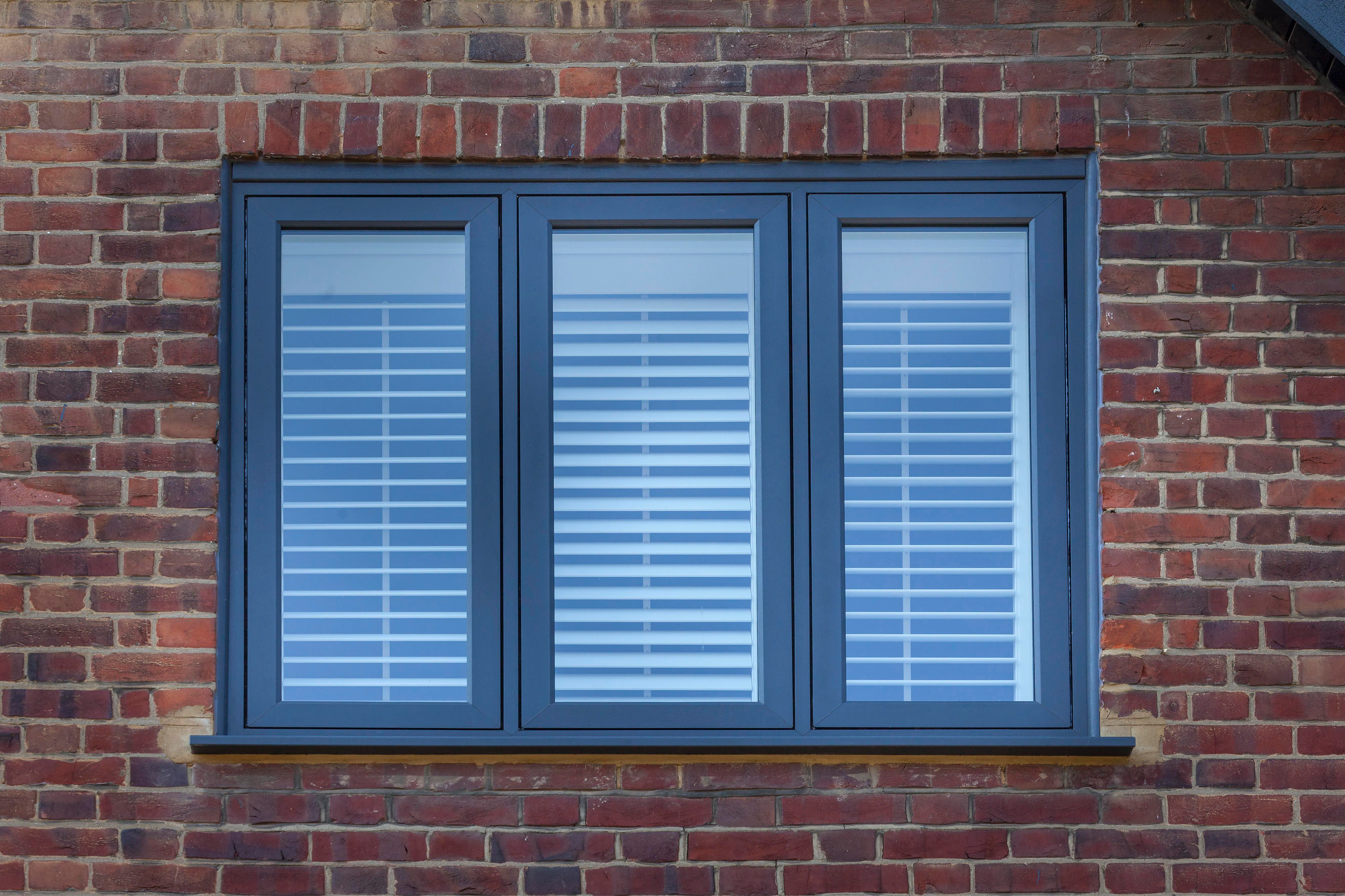 Three-panel window with blue frames set in a red brick wall, each panel having closed white horizontal blinds.
