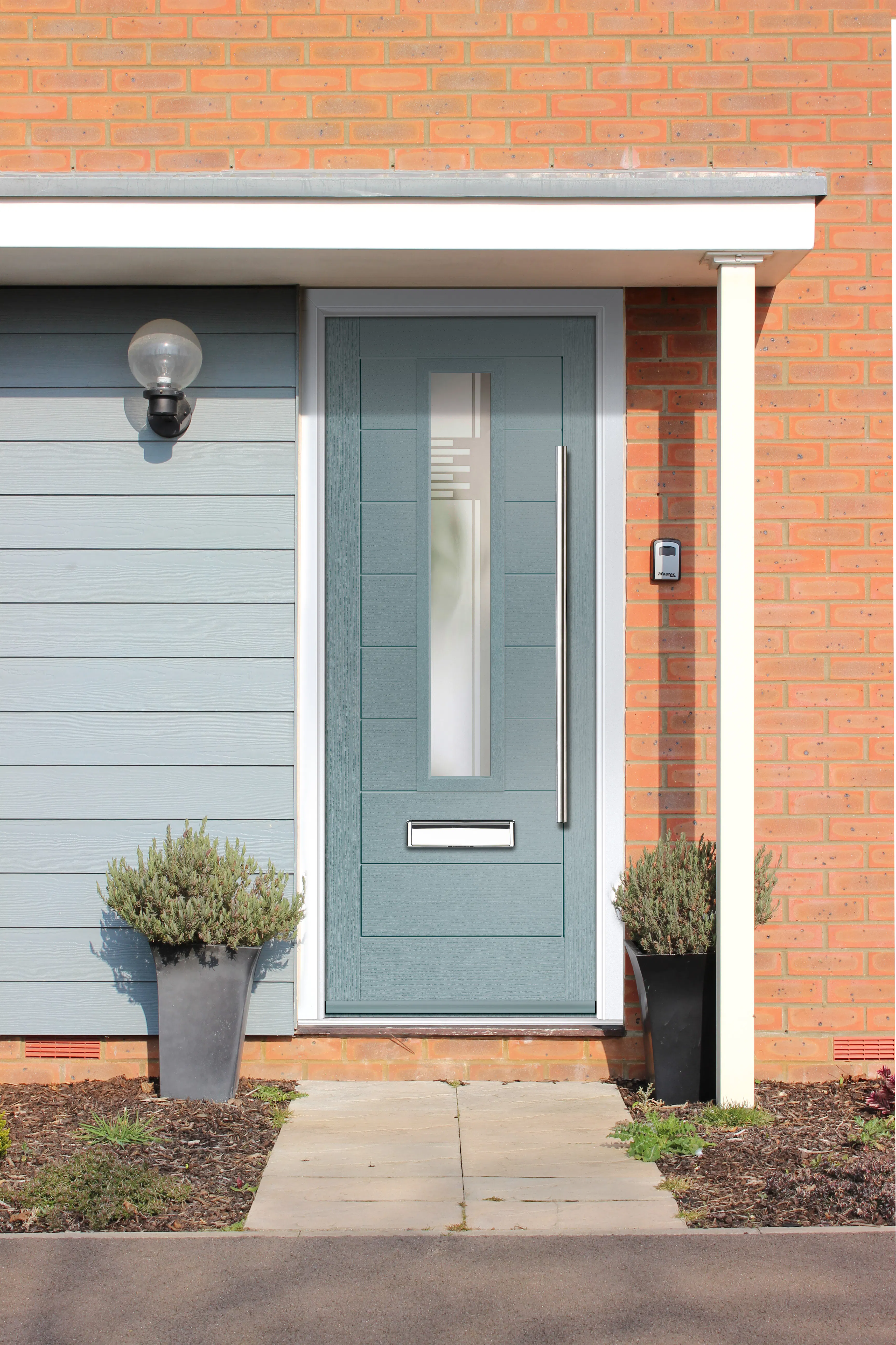 Front entrance of a house with a blue door featuring a vertical glass panel, silver handle, mail slot, and two black planters with green shrubs on either side.