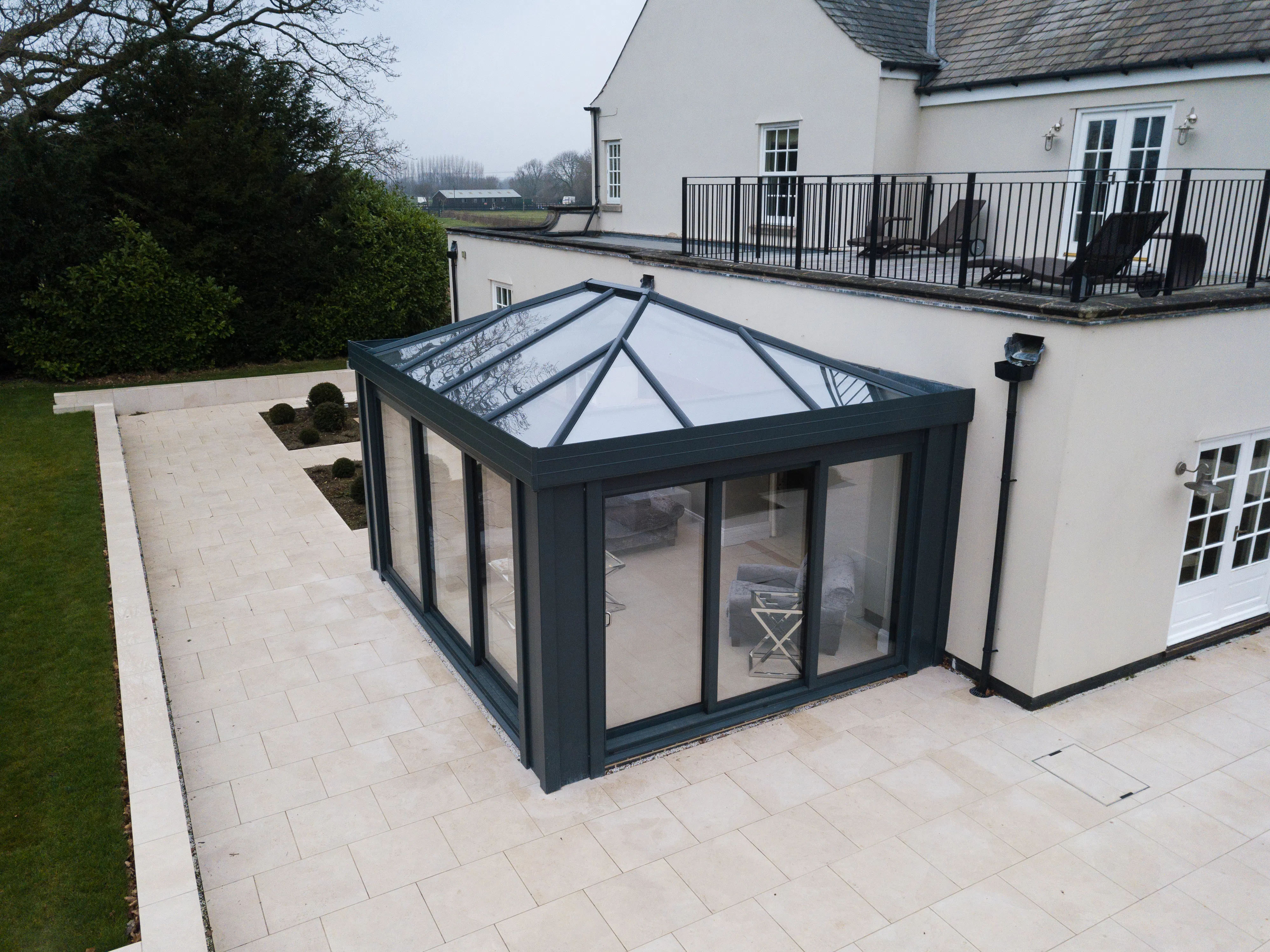 Single-story glass conservatory extension with dark framing attached to a beige house, surrounded by light tiled patio and garden area with shrubs.