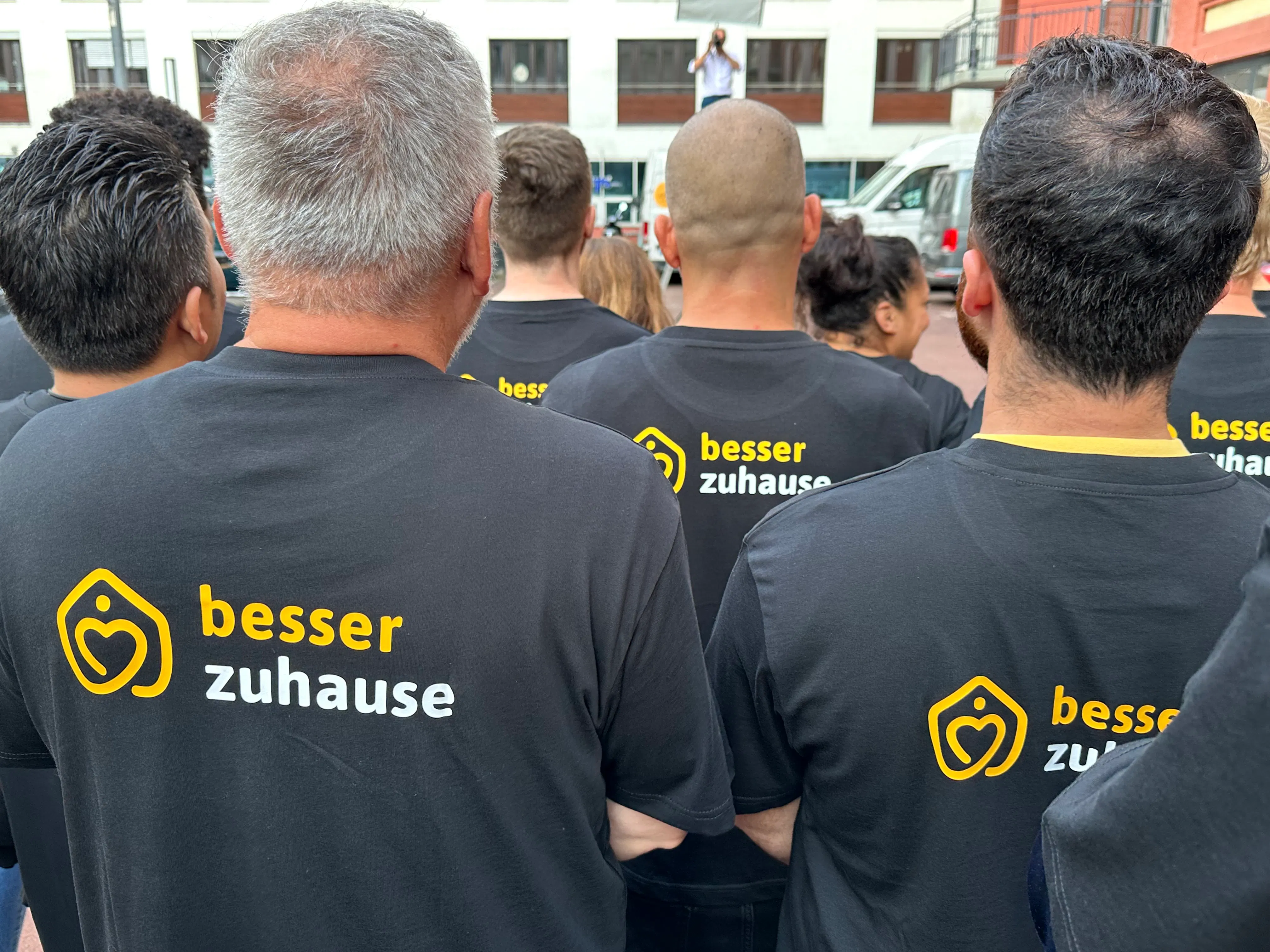 Menschen stehen mit dem Rücken zur Kamera, tragen schwarze T-Shirts mit dem gelben und weißen Schriftzug „besser zuhause“ und einem Herz-Symbol.